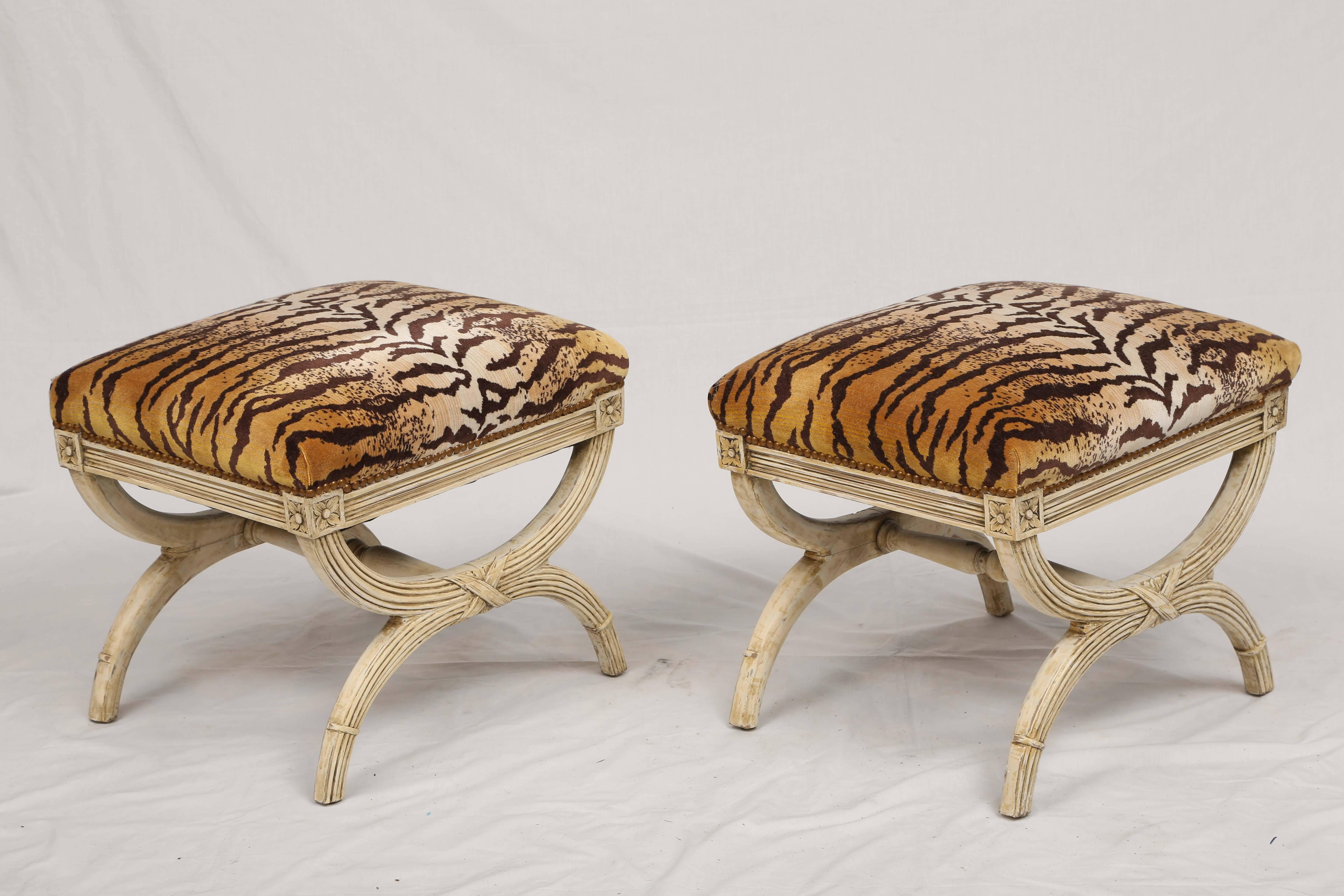Pair of stools, each having an crown seat in LeTigre silk velvet with nailheads, on painted X-frame, showing natural wear, its reeded seatrail flanked by rosettes, raised on tapering, curved legs, joined by crossed ribbon and turned stretcher.
