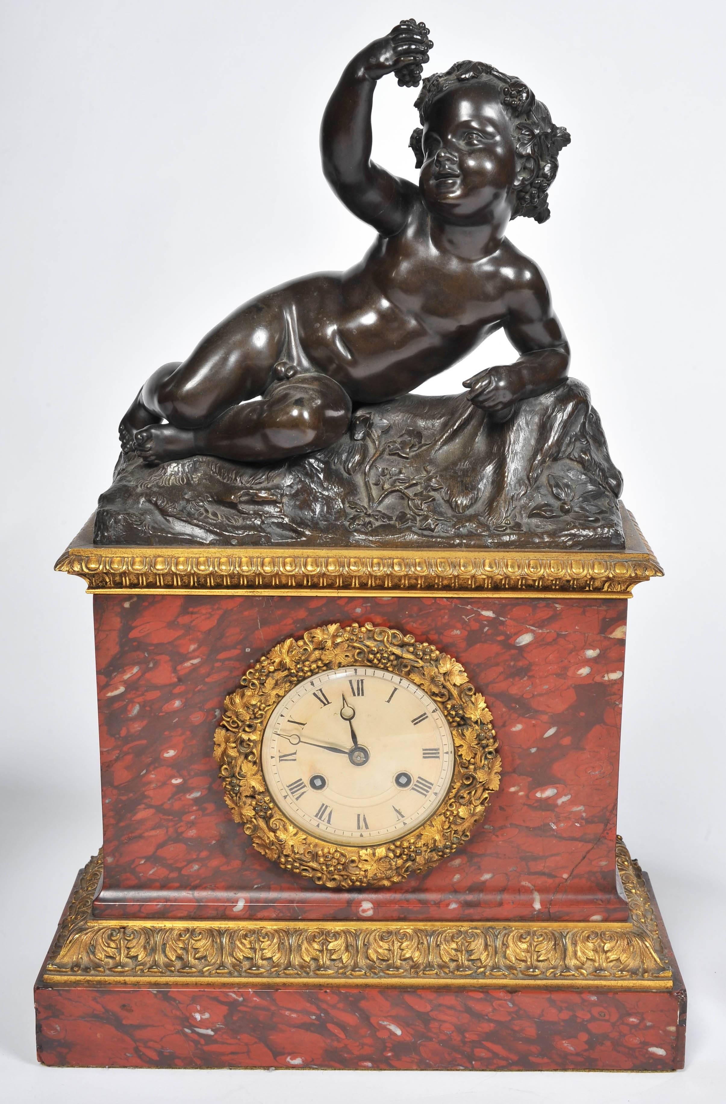 A very pleasing French 19th century rouge marble and gilded ormolu clock garniture. Having a bronze reclining putti holding a bunch of grapes. The silvered dial has an eight day bell chiming movement, the matching pair of bronze tazzas mounted on