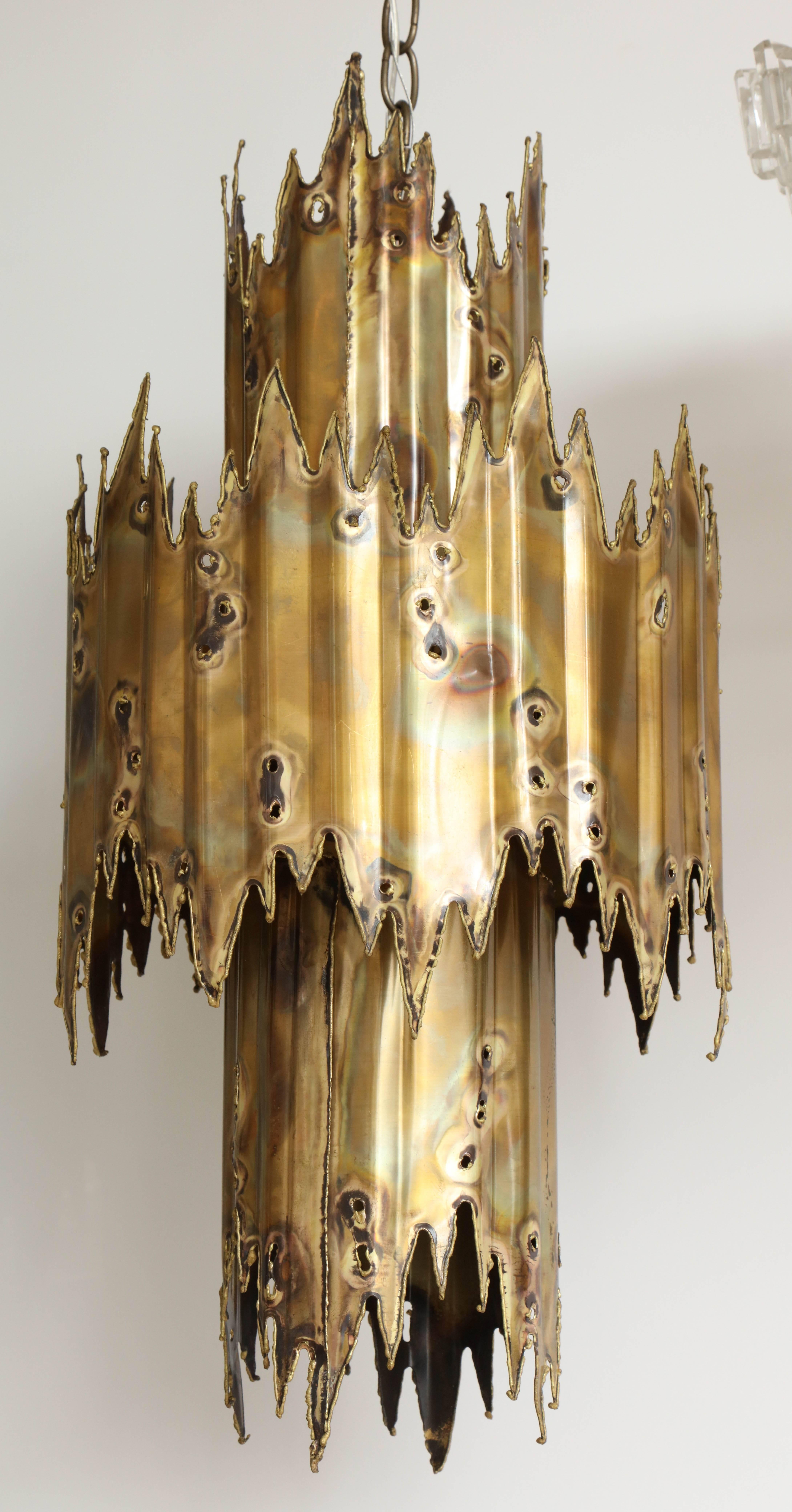 Mid-Century brutal style chandelier made from two layers of aged torch cut brass. Chandelier creates an amazing ambient glow when on. Suspended on an aged brass chain with canopy. Rewired for use in USA using three chandelier sockets and one center