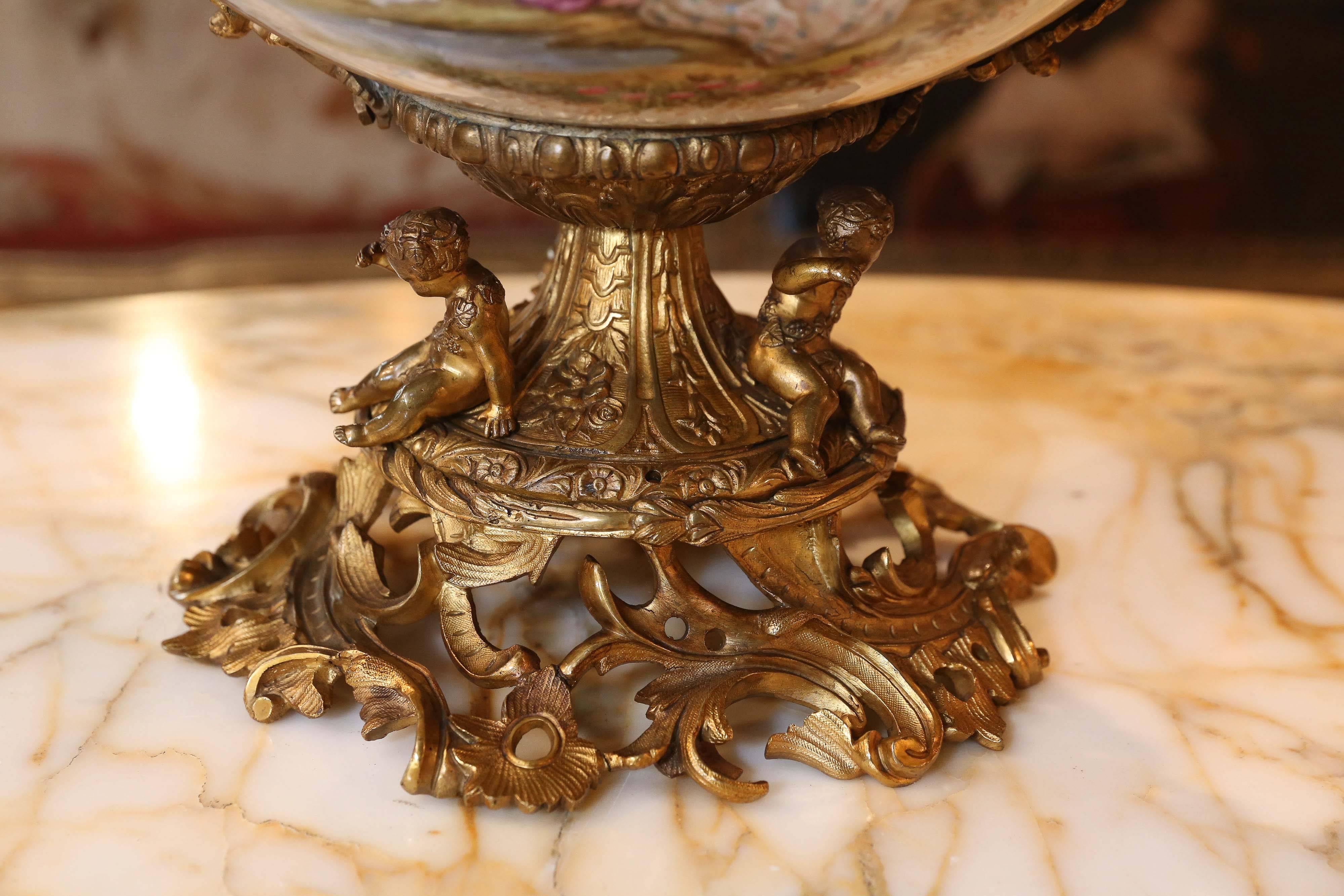 The top of this rim mounted with a wide pierced ormolu border and two 
scrolling handles, on a circular base with three putti on a scrolling pierced floral 
ormolu foot. The bowl with a Celeste blue ground and two large gilt framed panels of a