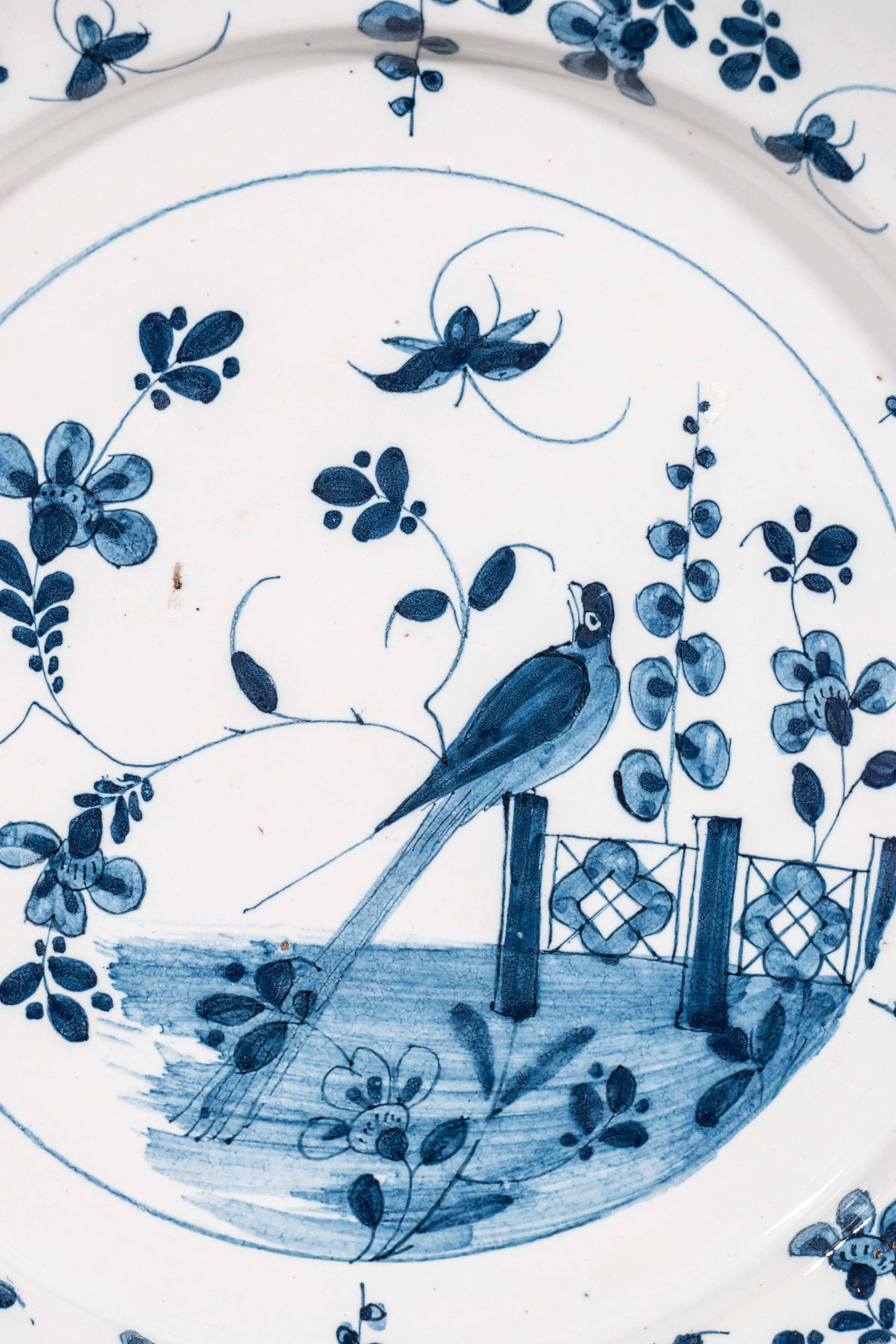 A Dutch Delft blue and white charger showing a lively garden scene with a butterfly and a bird sitting on the garden fence. The border decorated with smaller butterflies, flowers and scrolling vines.
    