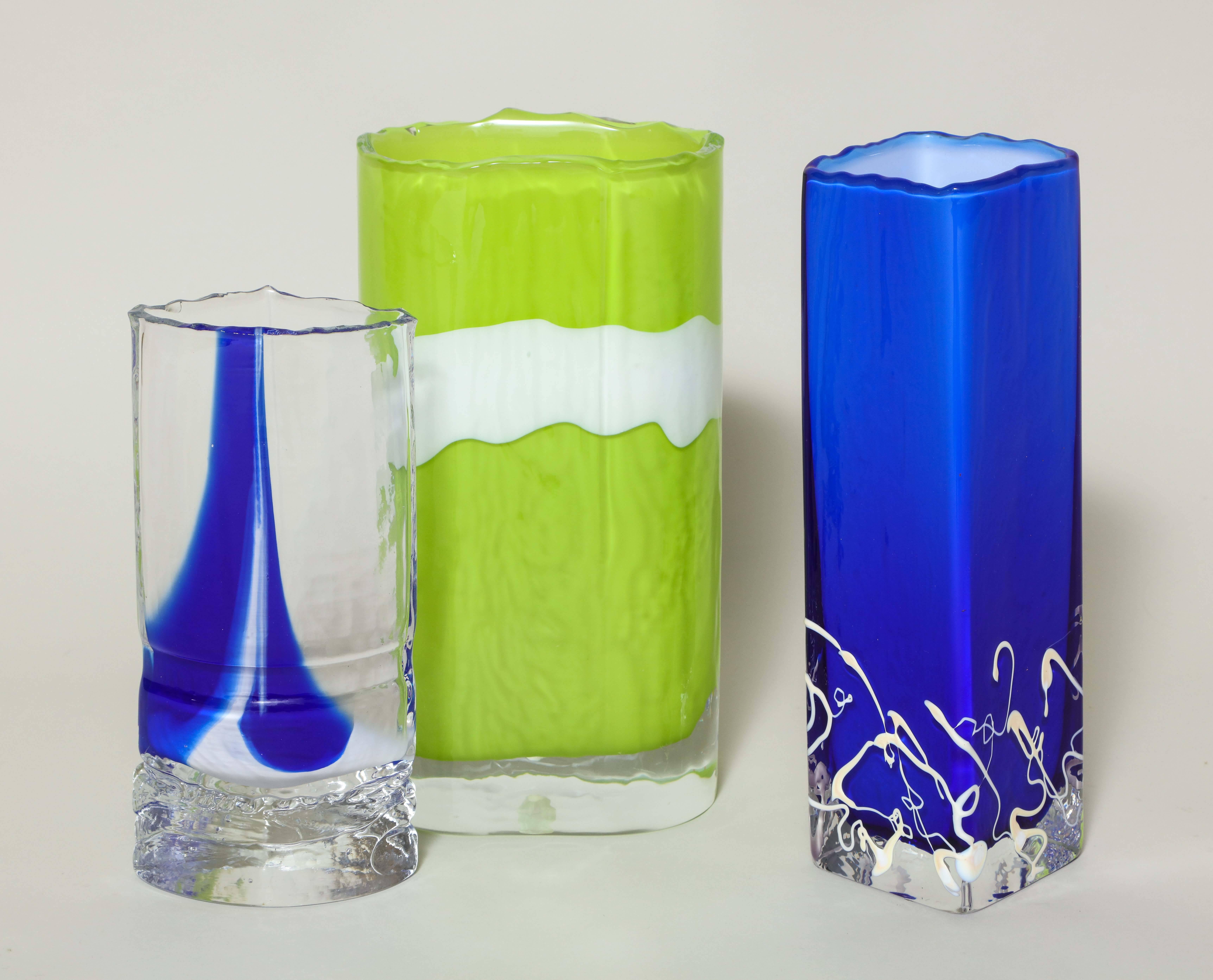 A collection of three vibrantly hued handmade sculptural glass vessels by Berànek with reticulated top edges and vibrant colors with white accents: lime green with white ribbon, blue with white drip lines and blue with white inclusion. Emanuel