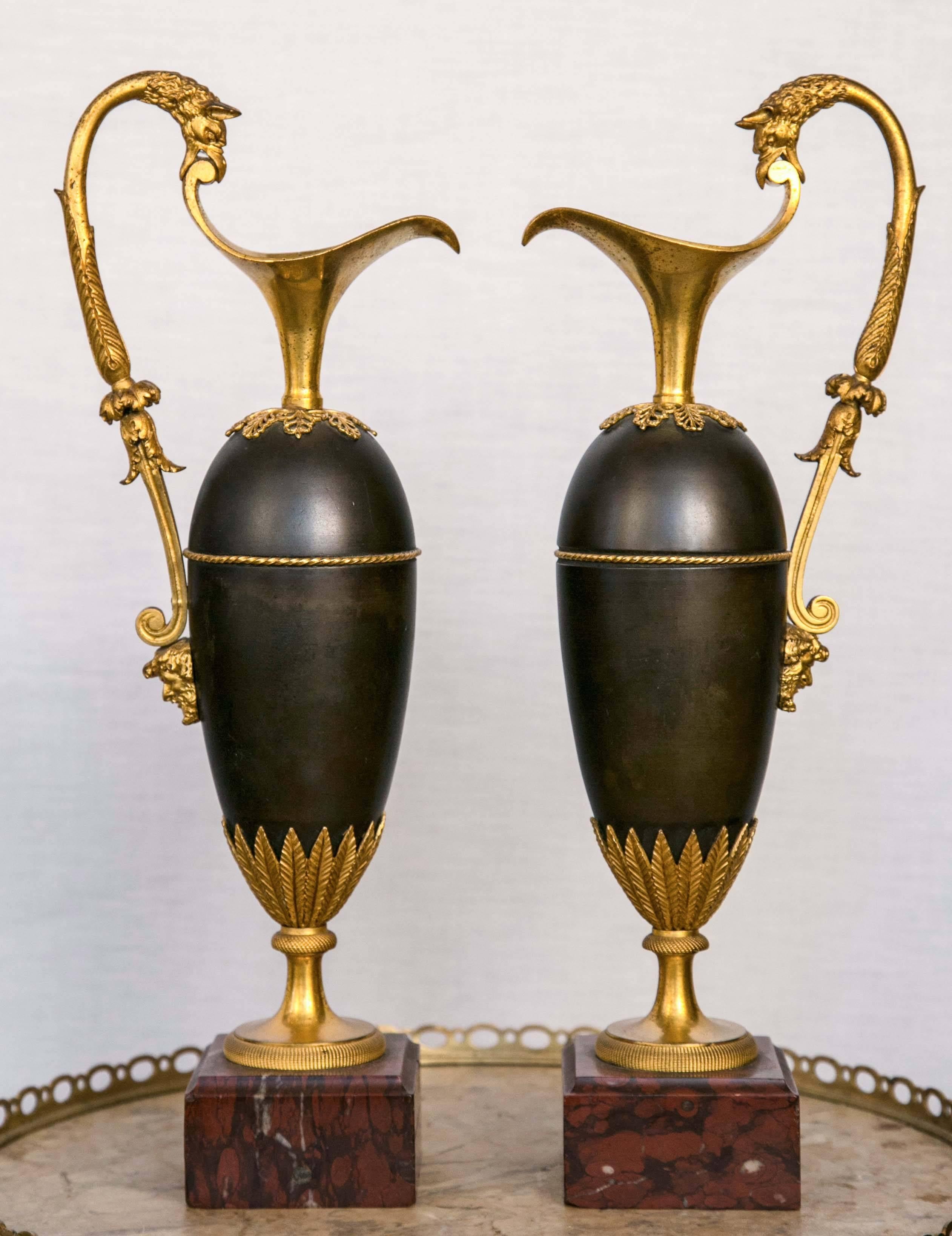 A superb pair of bronze ewers from the French Empire period with gilt bronze swooping high handles ending in horned bird heads attached to the spouts, below which are bearded male masks. Open work palmettes below the spouts, a ring of tightly