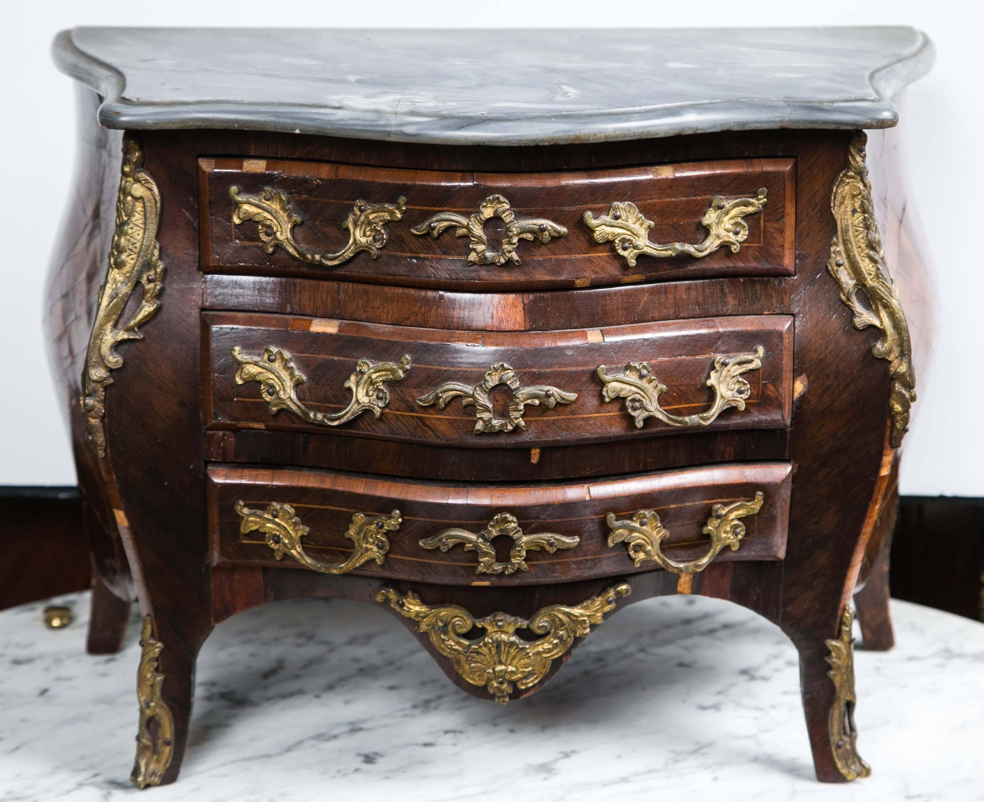 A salesman's sample miniature chest with a replaced grey marble chest. The marble glued onto the top.
Three drawers with bronze pulls and mounts, bronze mounts on the shaped body, apron and front feet. 
Thick veneers in parquetry patterns. Some