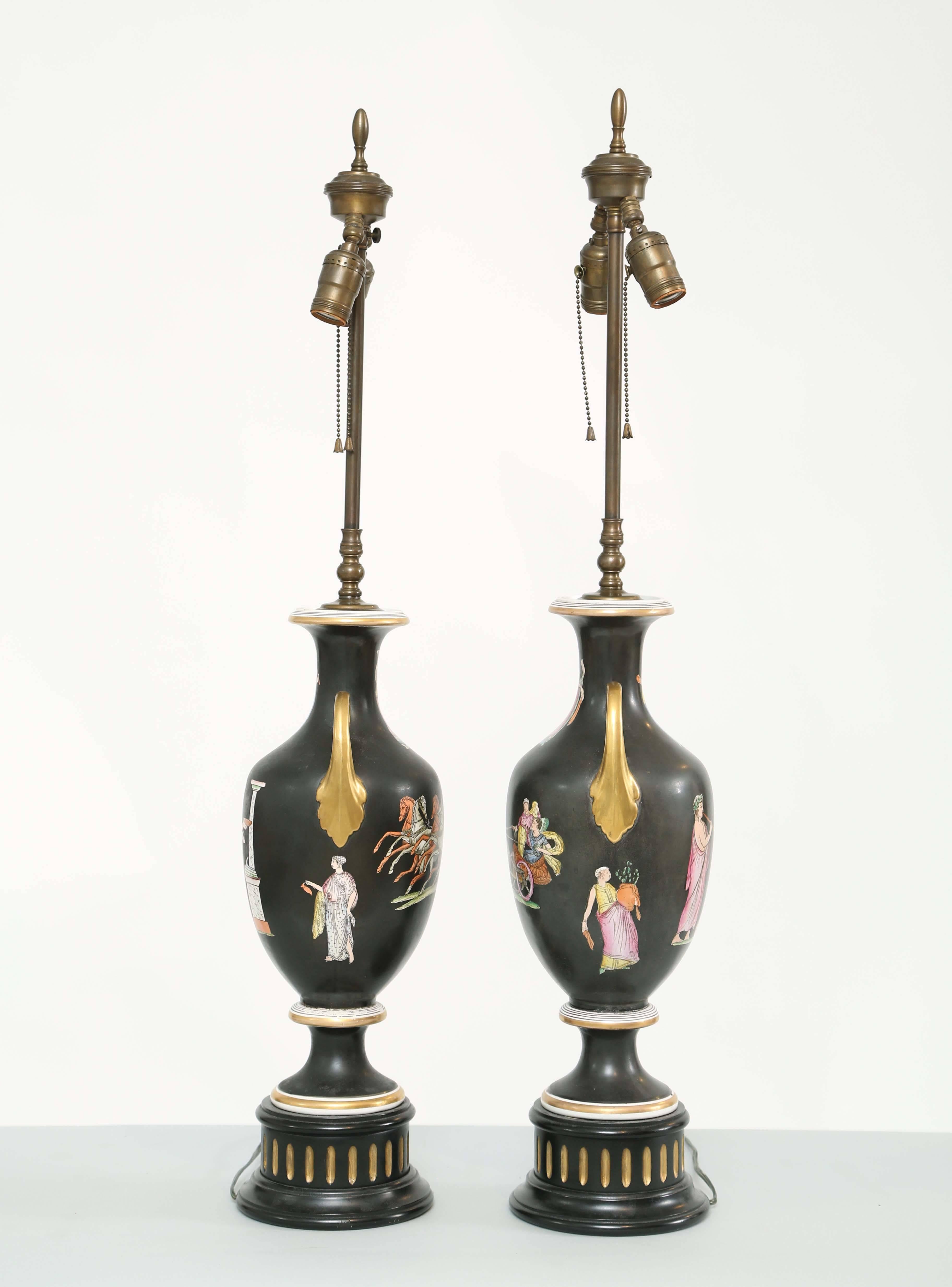 Pair of black Amphora urn-form porcelain vases, each with colorful transfer decoration of Charioteers and Classical figural motifs, having a flared neck flanked by gilded handles, raised on round foot and fluted plinth.