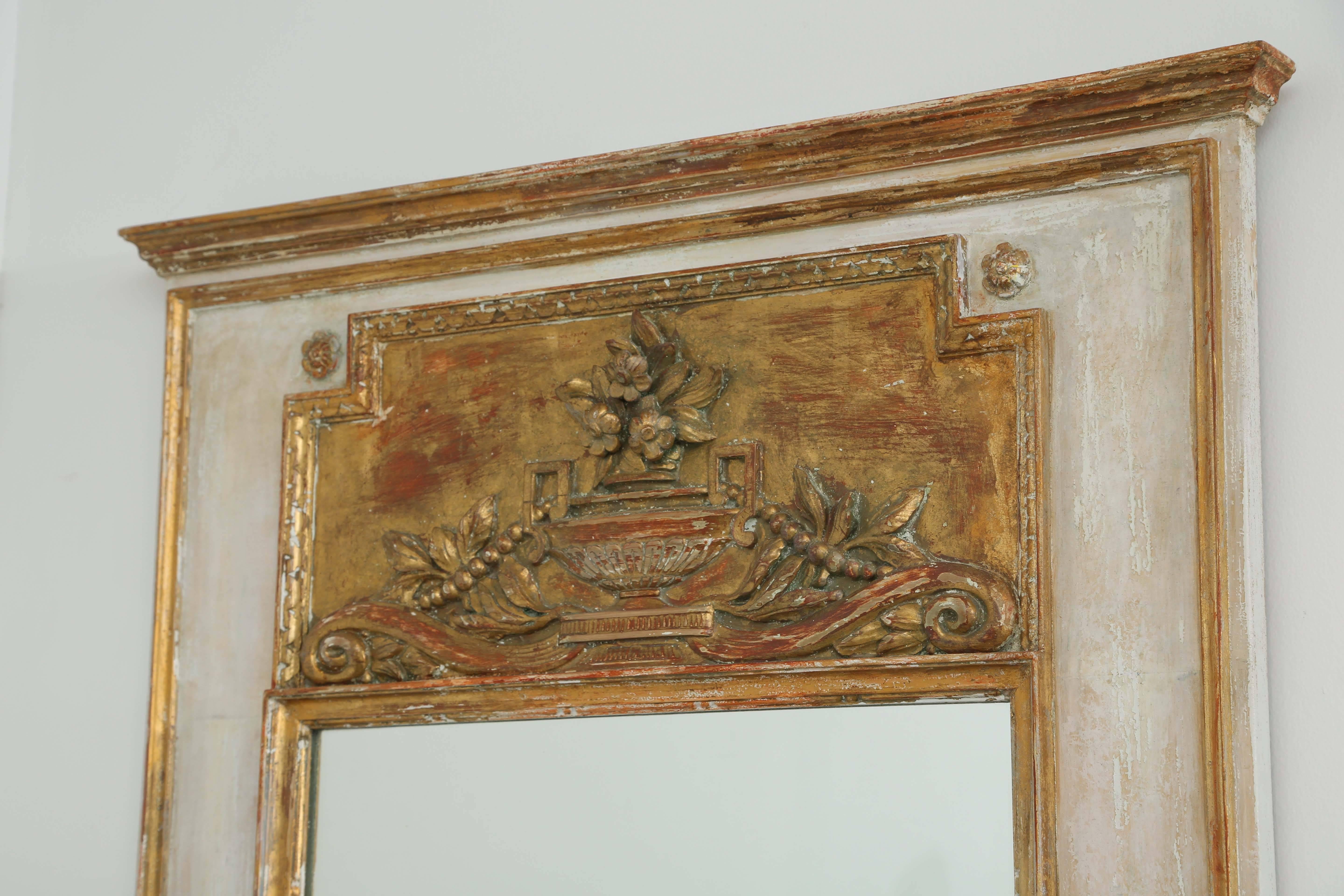 Trumeau mirror, with painted and parcel-gilt finish, having a stepped cornice, over decorative panel outcarved with a flower-filled urn, flanked by acanthus and scrolls, in gadrooned border, surmounting its rectangular looking glass in molded