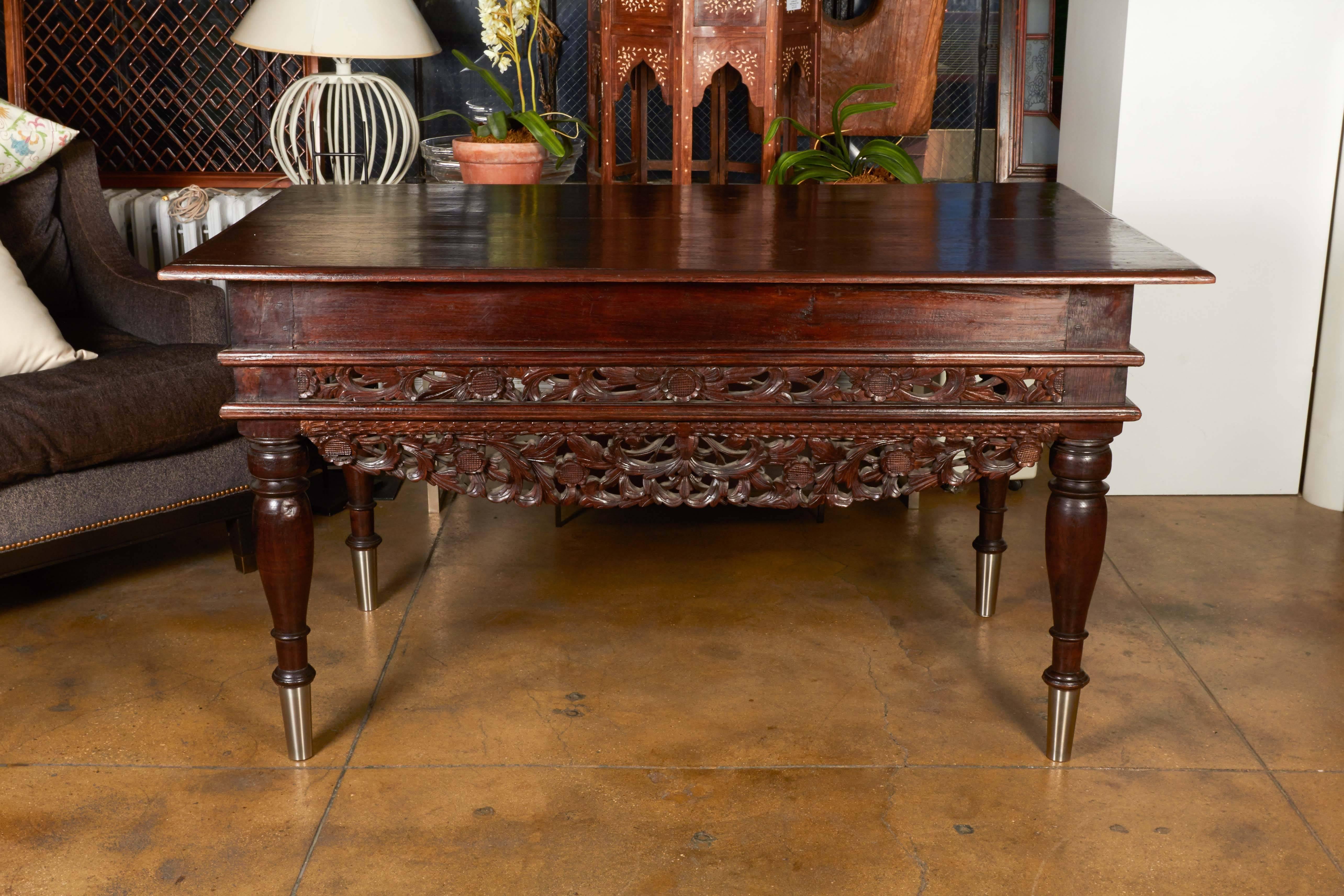 A desk or table from Madura, an island in Indonesia, with an ornate floral carving on its front and apron, scrolled legs with stainless feet, and one small drawer.