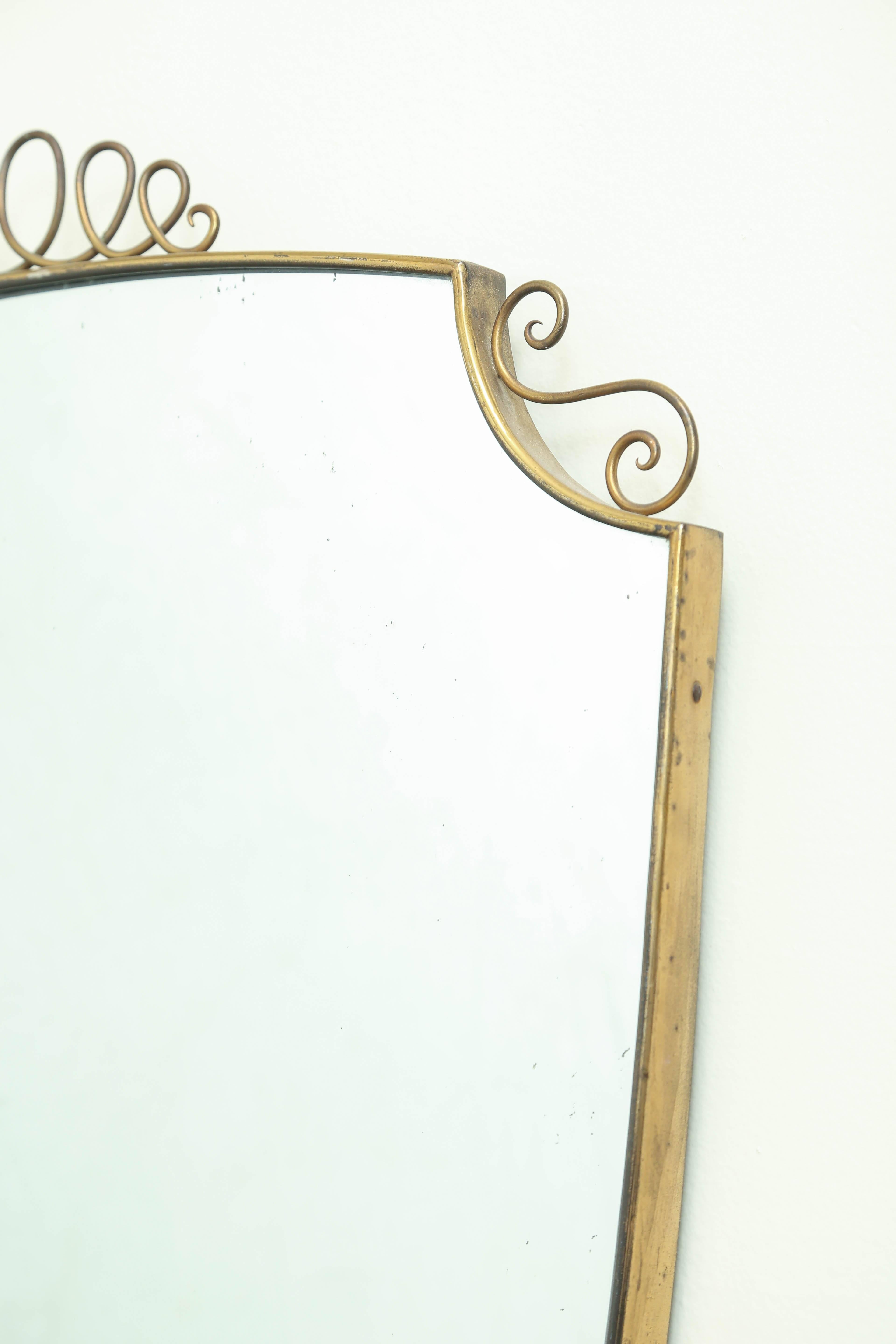 A large 1950s, Italian shield shape brass frame mirror in the style of Gio Ponti.
The frame is handmade with top multi loop and side scroll accents.