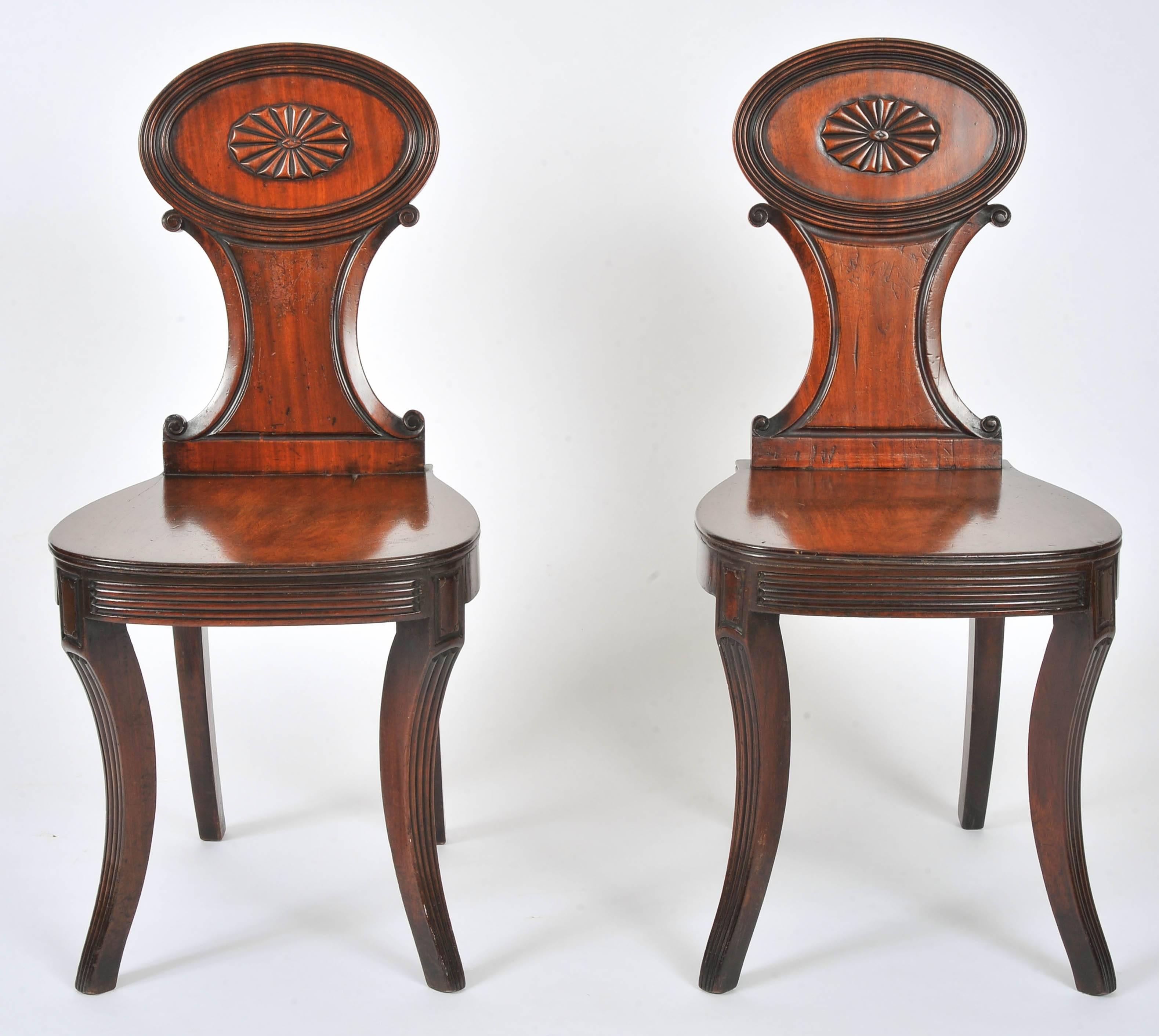 This lovely pair of 19th century. Carved mahogany hall chairs features a shaped back splat that has a decorative oval top section with a medallion centrepiece. The chairs feature sabre front and back swept legs and hide shaped seats. Each chair