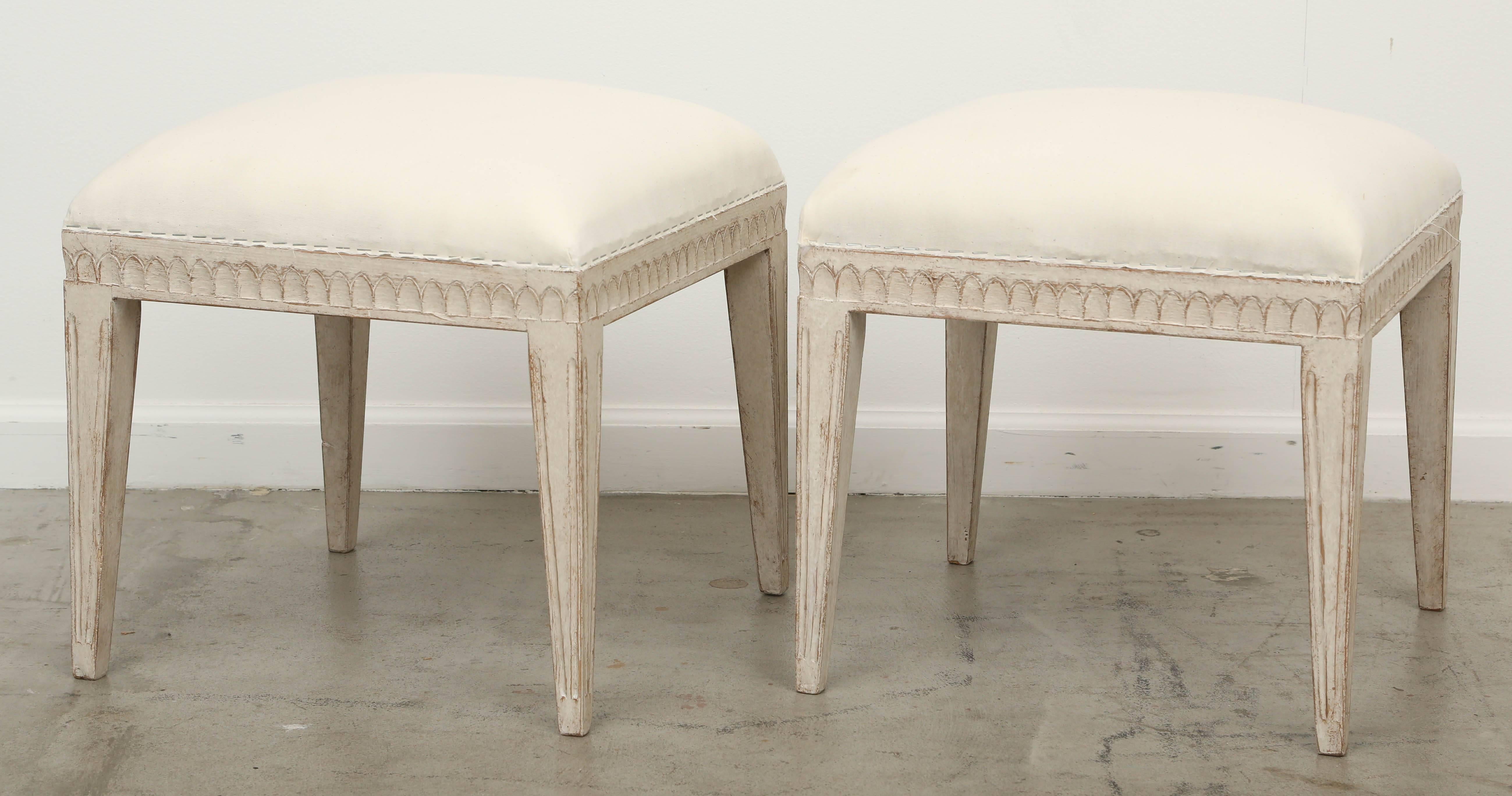 Pair of Swedish Gustavian painted stools, mid-19th century. 
Carved lambs tongue border on apron, squared tapered legs, stools are painted in grayish white distressed finish and upholstered seats. 

Measures: 18 in W x 18 in D x 17 in H.