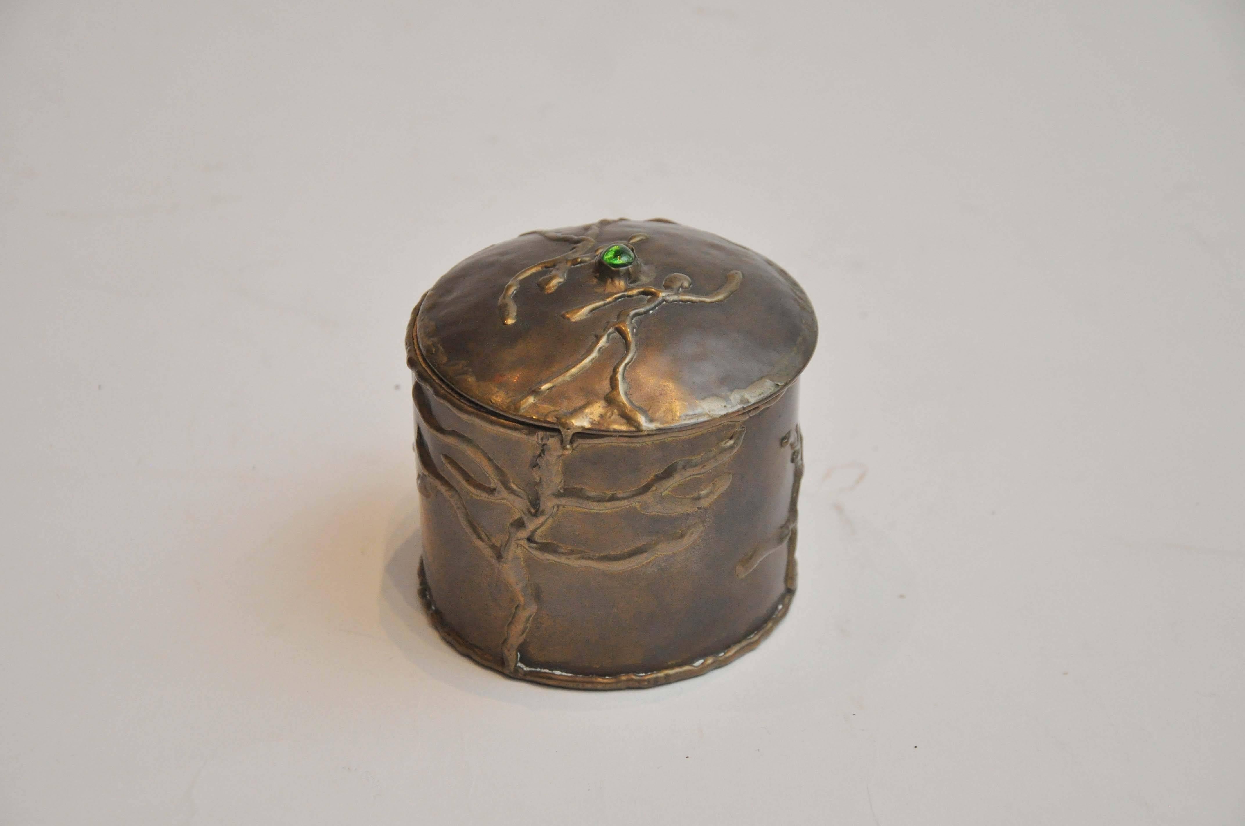 Early 20th century round brass box with cabochon stone by artist R Cervantes. Beautiful box with hinged lid. Features a raised branch detail and an emerald cabochon stone adorns the lid of the box.

Dimensions: 4"Diam x 3.75"H.