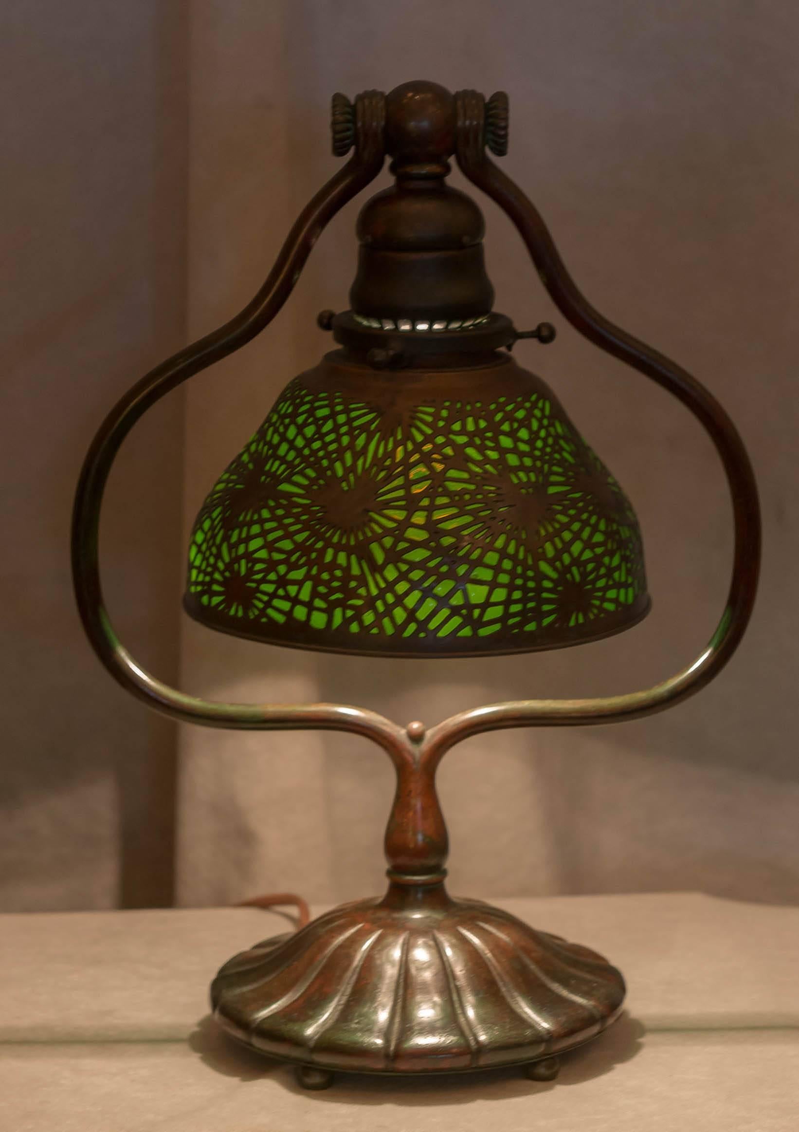 This sweet little lamp is a nice example of the work of the master of lamps, Tiffany Studios. The bronze base has that rich patina, and the shade is green glass encased in the pine needle design.The base is signed. These shades were never signed,