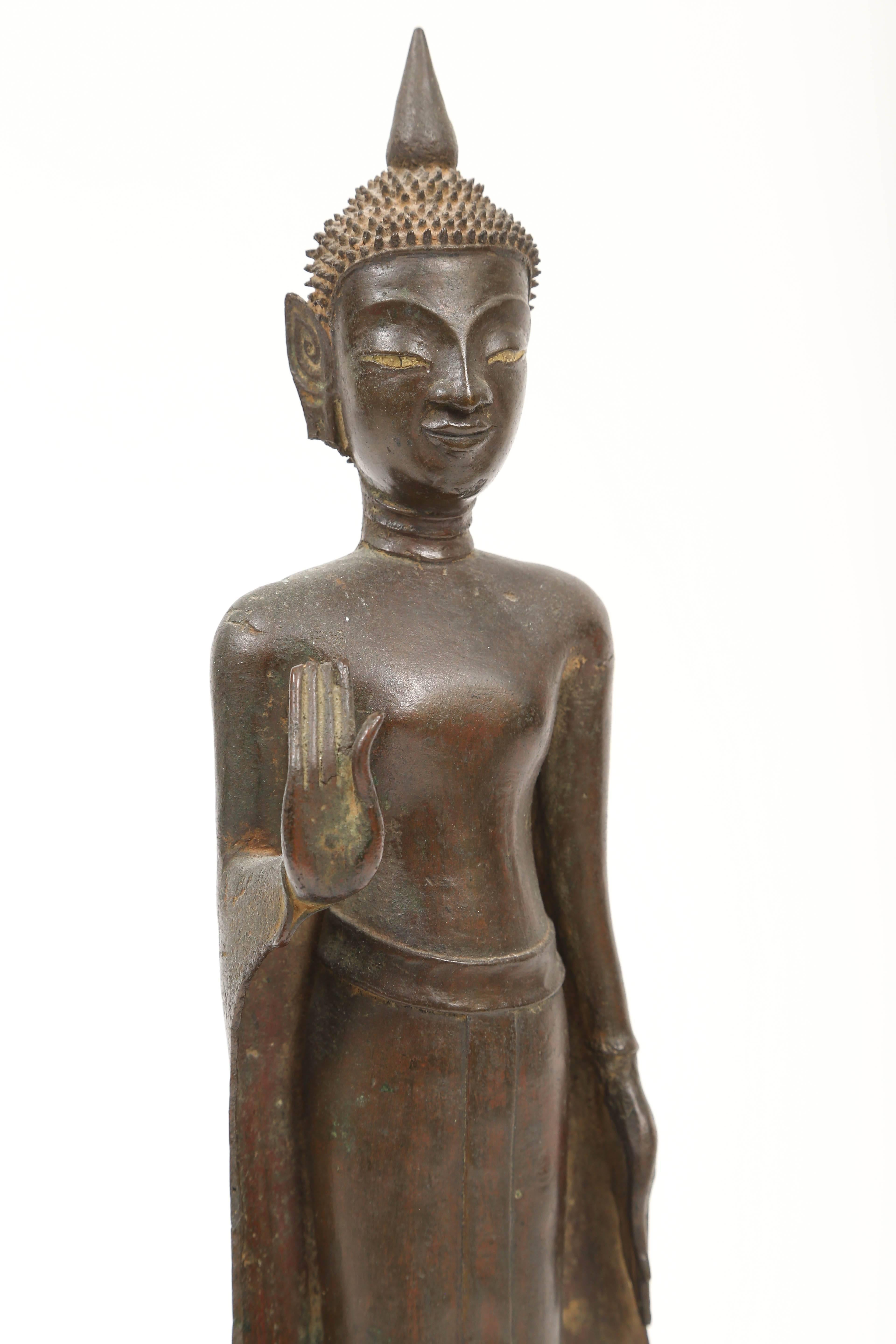 This bronze standing Buddha mounted on a turned wood base has his right palm held outward in the abhaya mudra position, a gesture of fearlessness and reassurance.