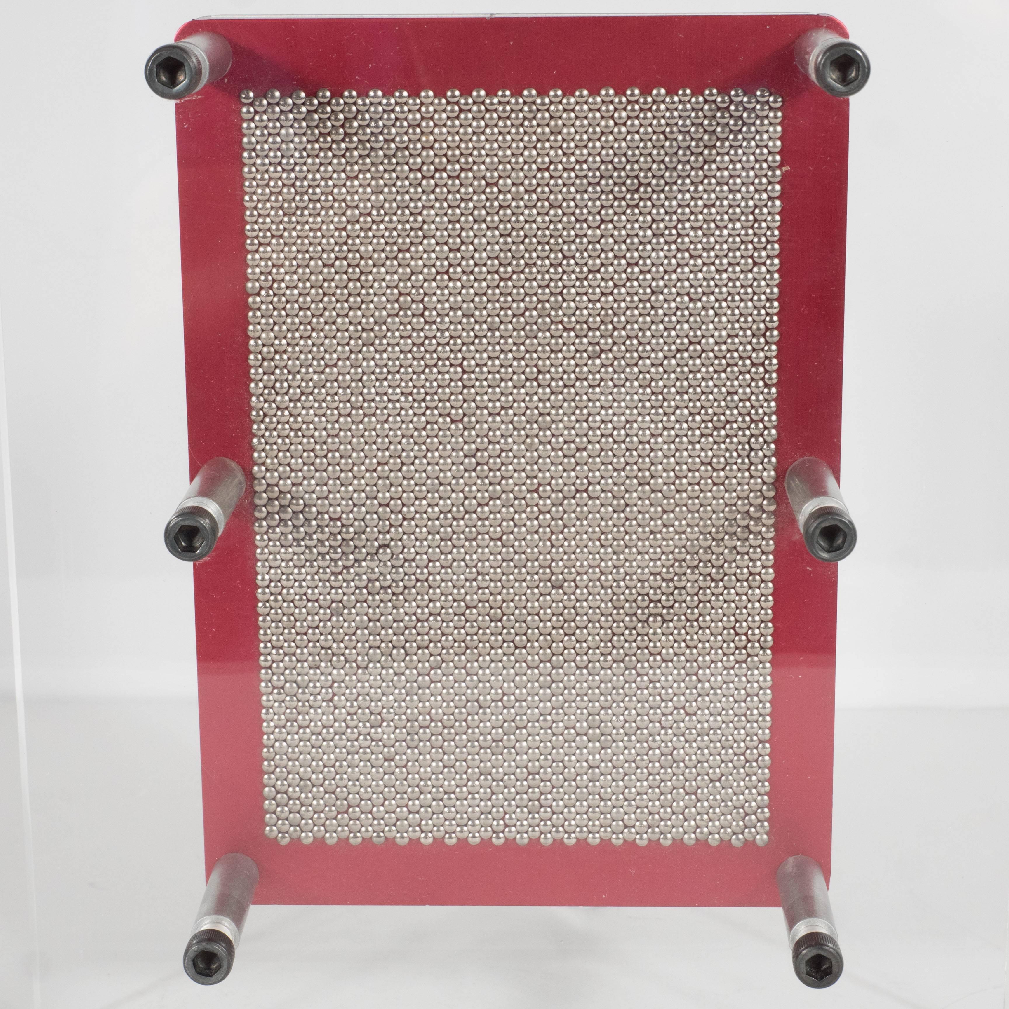 A Mid-Century pin-point impression box with thick Lucite frame. Two rectangular Lucite frames support a central, floating red steel mounted plaque which holds thin, flathead nails. A temporary impression is made by pressing on the narrow protruding
