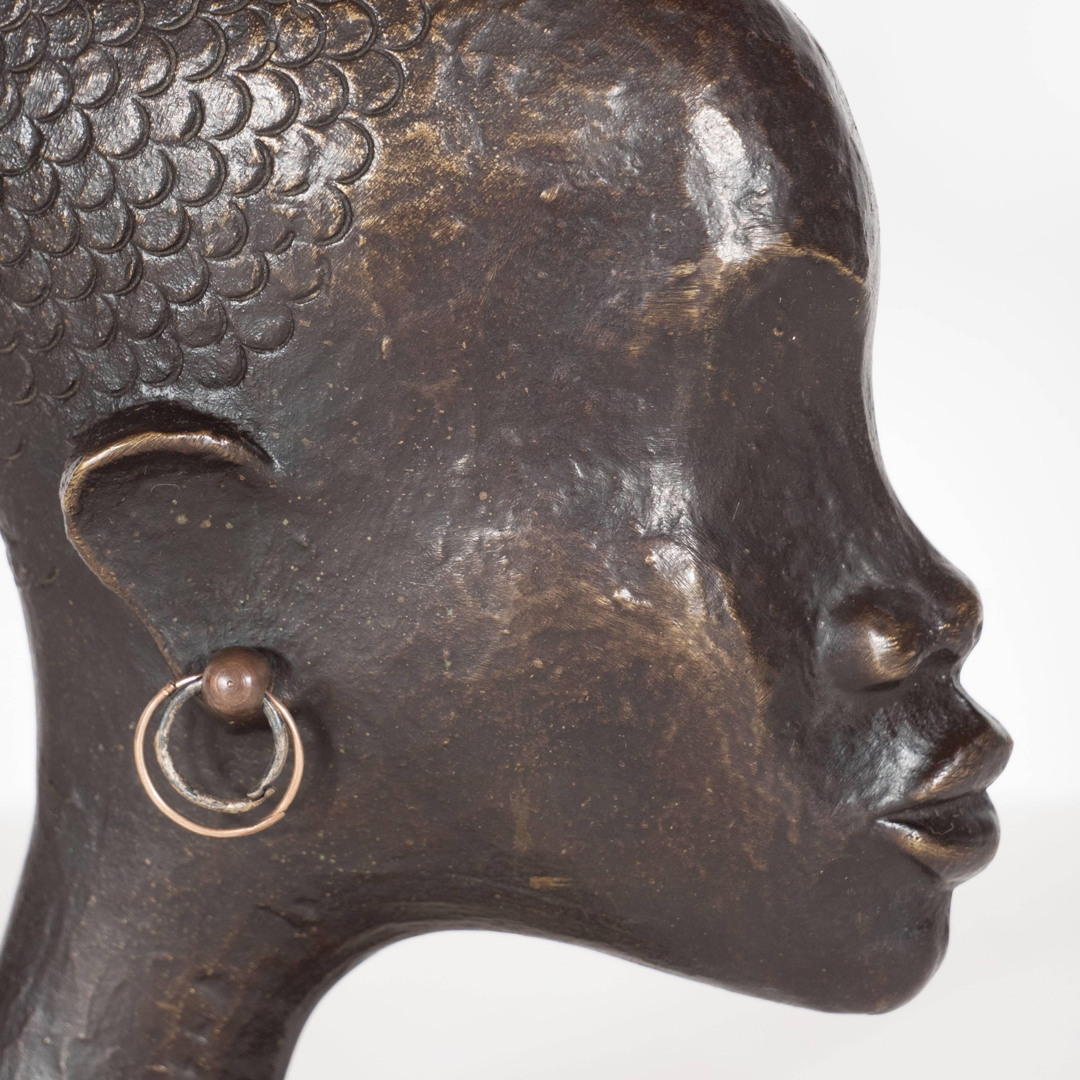 A Mid-Century Modernist patinated bronze of an African woman's head in profile by Werkstatte Hagenauer. She is adorned with brass hoop earrings. In place of hair are fish scales in the form of a splayed tail. A playful yet elegant example of