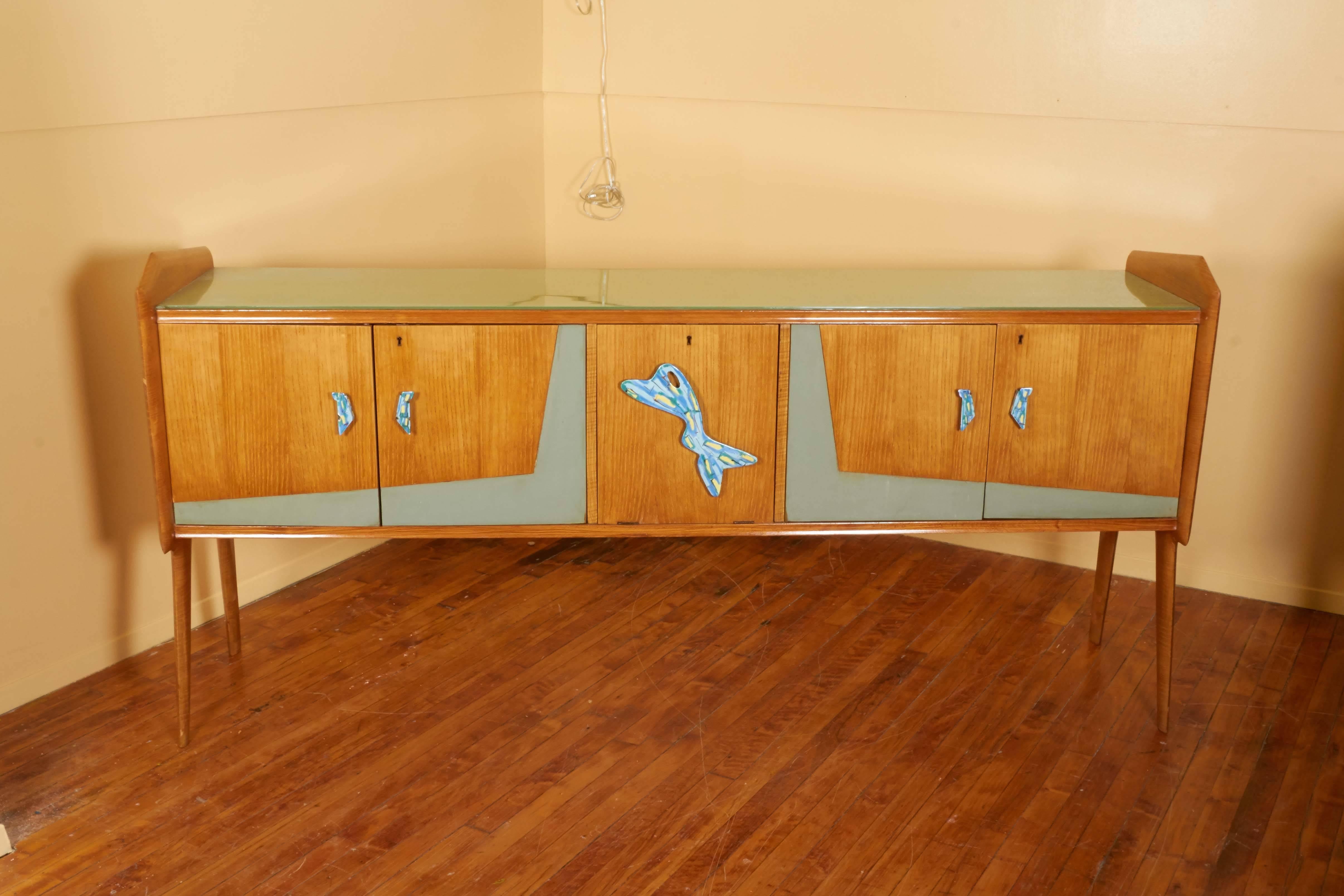 A lengthy circa 1950s sideboard, produced in Italy, with glass top on wood frame, sides raised on tapered and slanted wood legs, including three cabinets for storage space, each with decorative biomorphic pull handles with enamel finish. Excellent