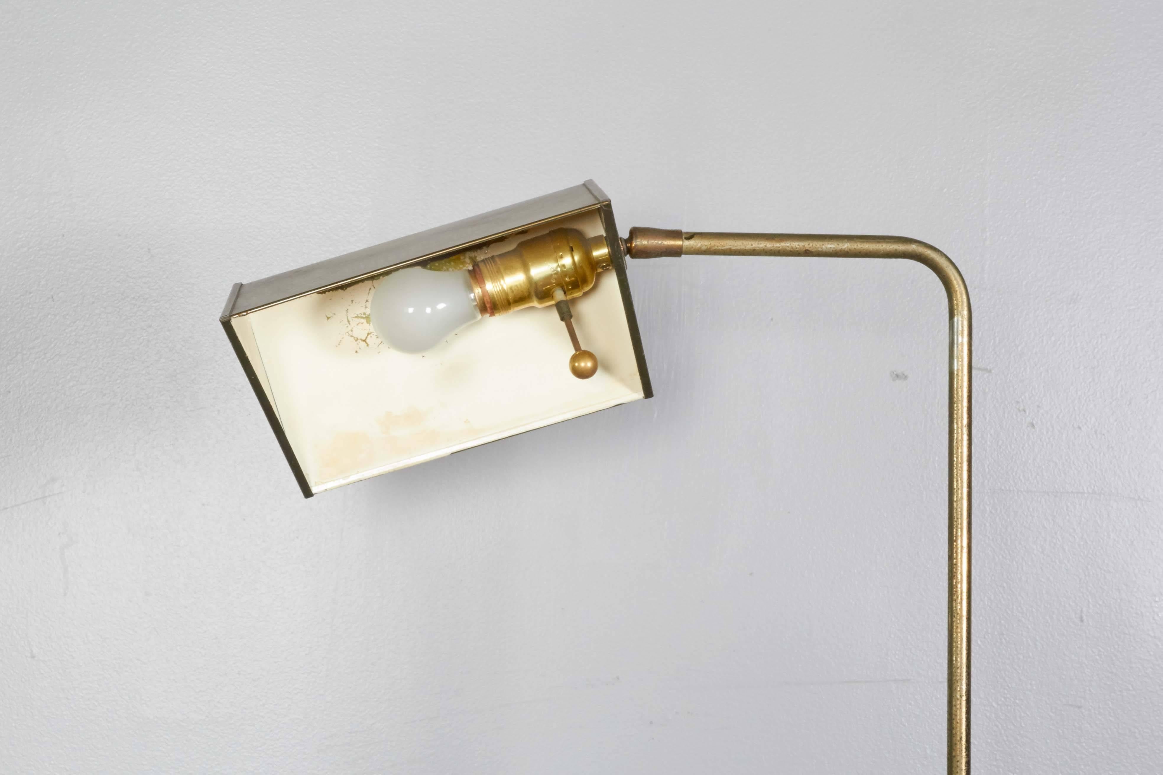 A brass circa 1960s pharmacy floor lamp, tent form shade with trimmed border and white enamel interior, on adjustable curved stem and round base. Good vintage condition, with age appropriate patina and wear to enamel interior, scratch to base.