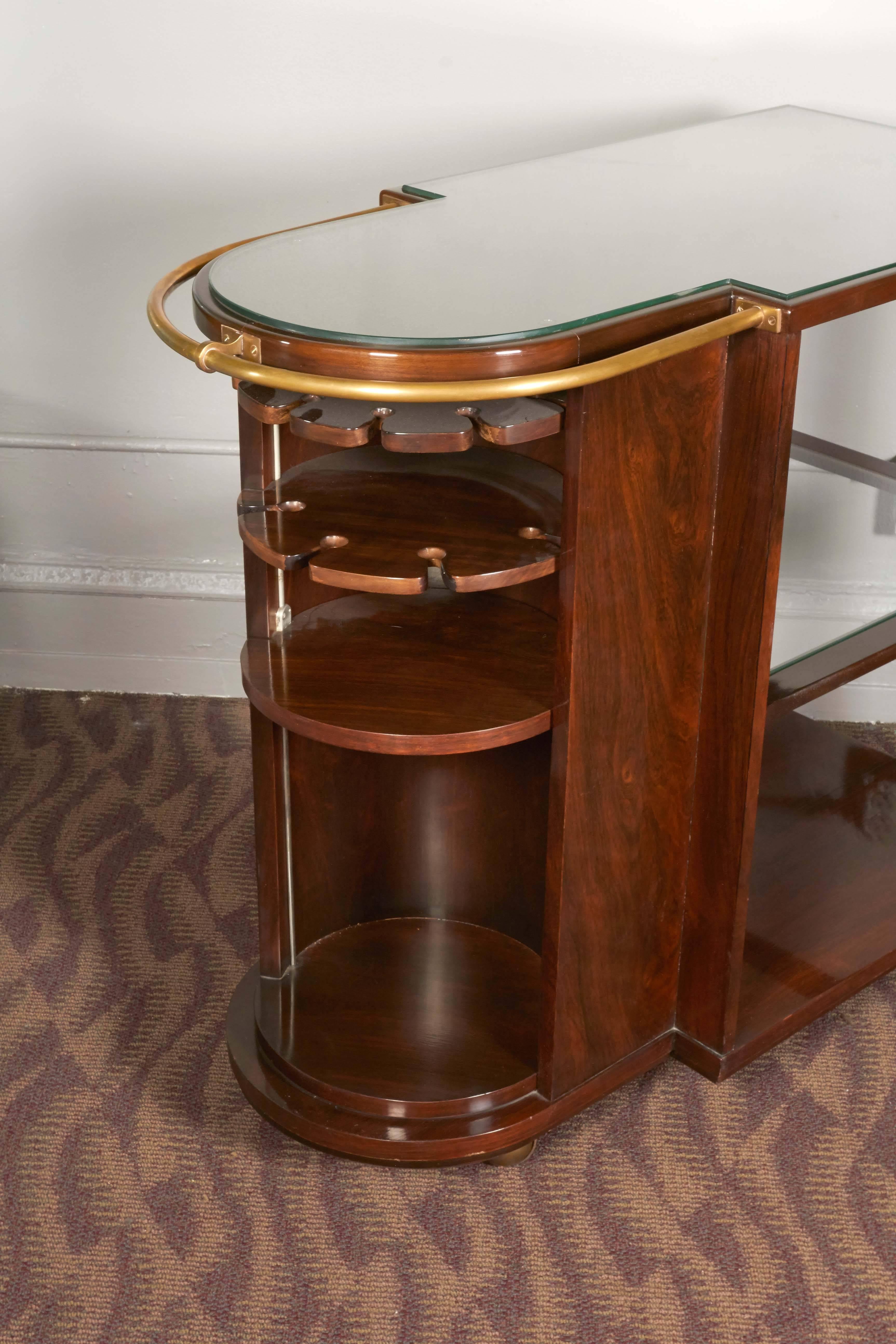 An Art Deco cocktail bar cart, produced circa 1940s, in mahogany with high gloss finish, accented with brass handle on caster wheels, including lower shelf and featuring a concealed cabinet with racks and storage space. Very good vintage condition