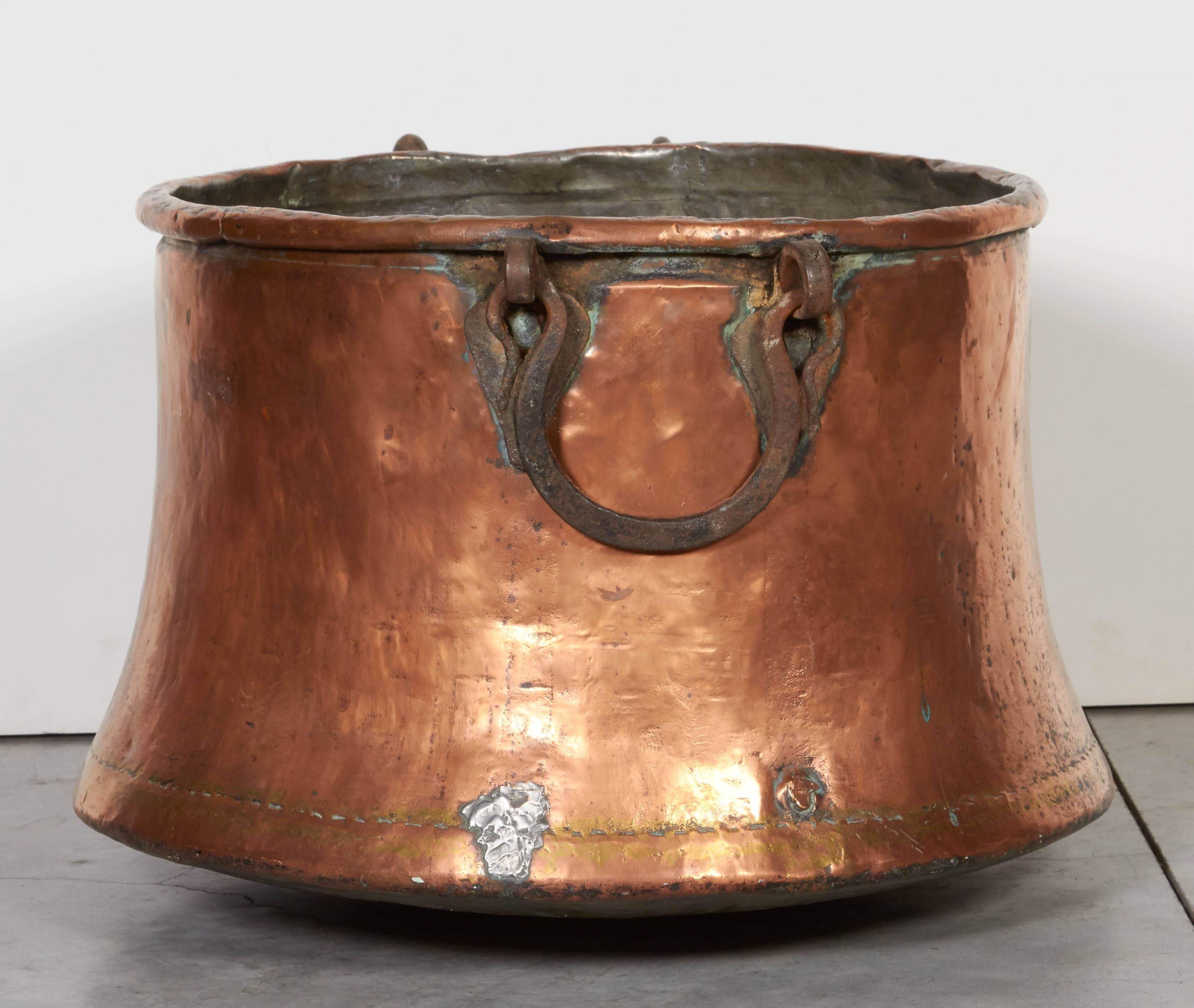 A large antique, tin lined joined copper kettle with a graceful flared base and beautifully hand-forged heavy iron handles. This striking pot has an unusually beautiful patina featuring several old repairs. A wonderful rustic vessel for firewood,