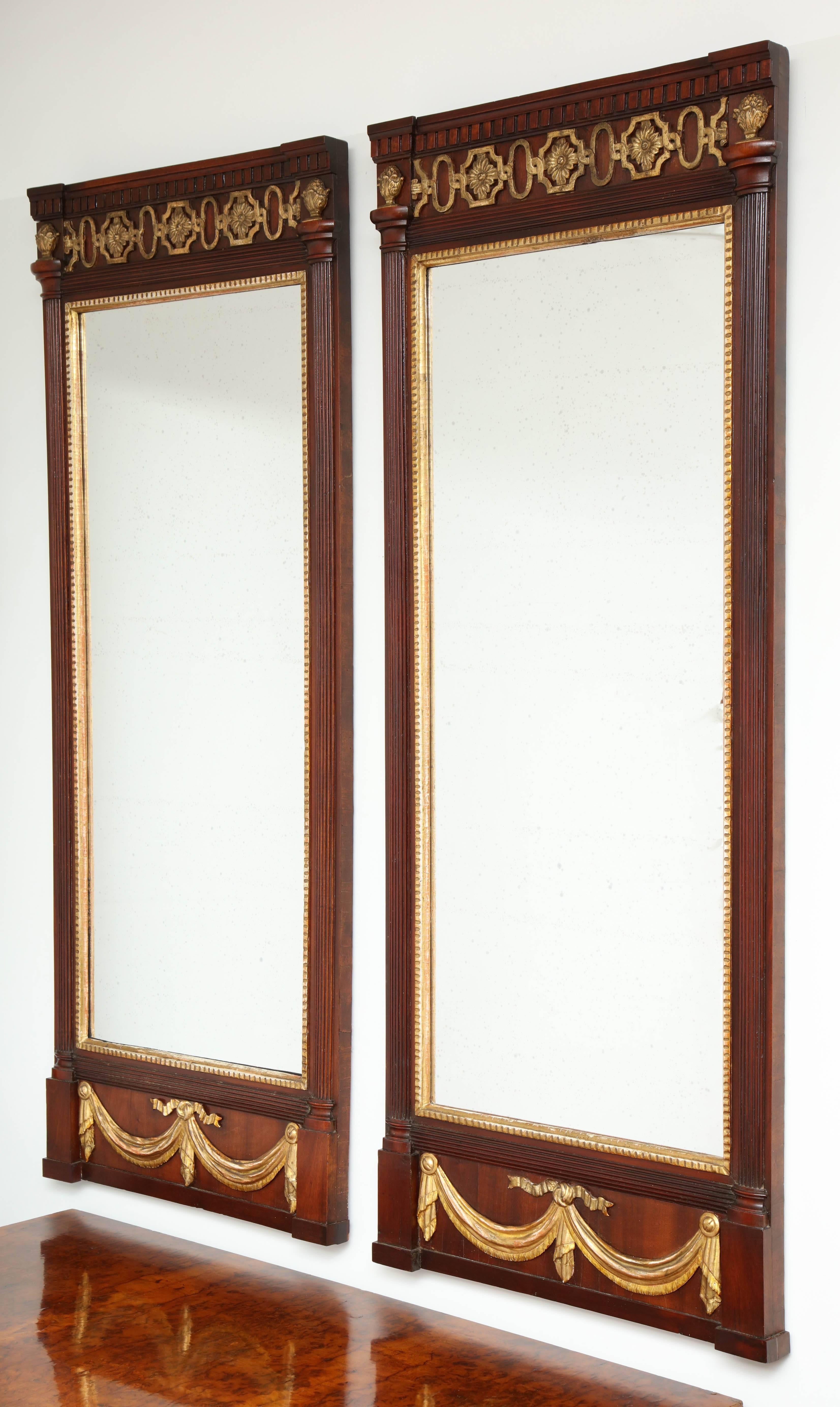 A pair of Danish Louis XVI mahogany and parcel-gilt mirrors, circa 1790s, each with a dentil frieze, half round pilaster and a gilded drapery swag. Both mirrors have replaced restoration mirror glass. These mirrors were probably set in a paneled