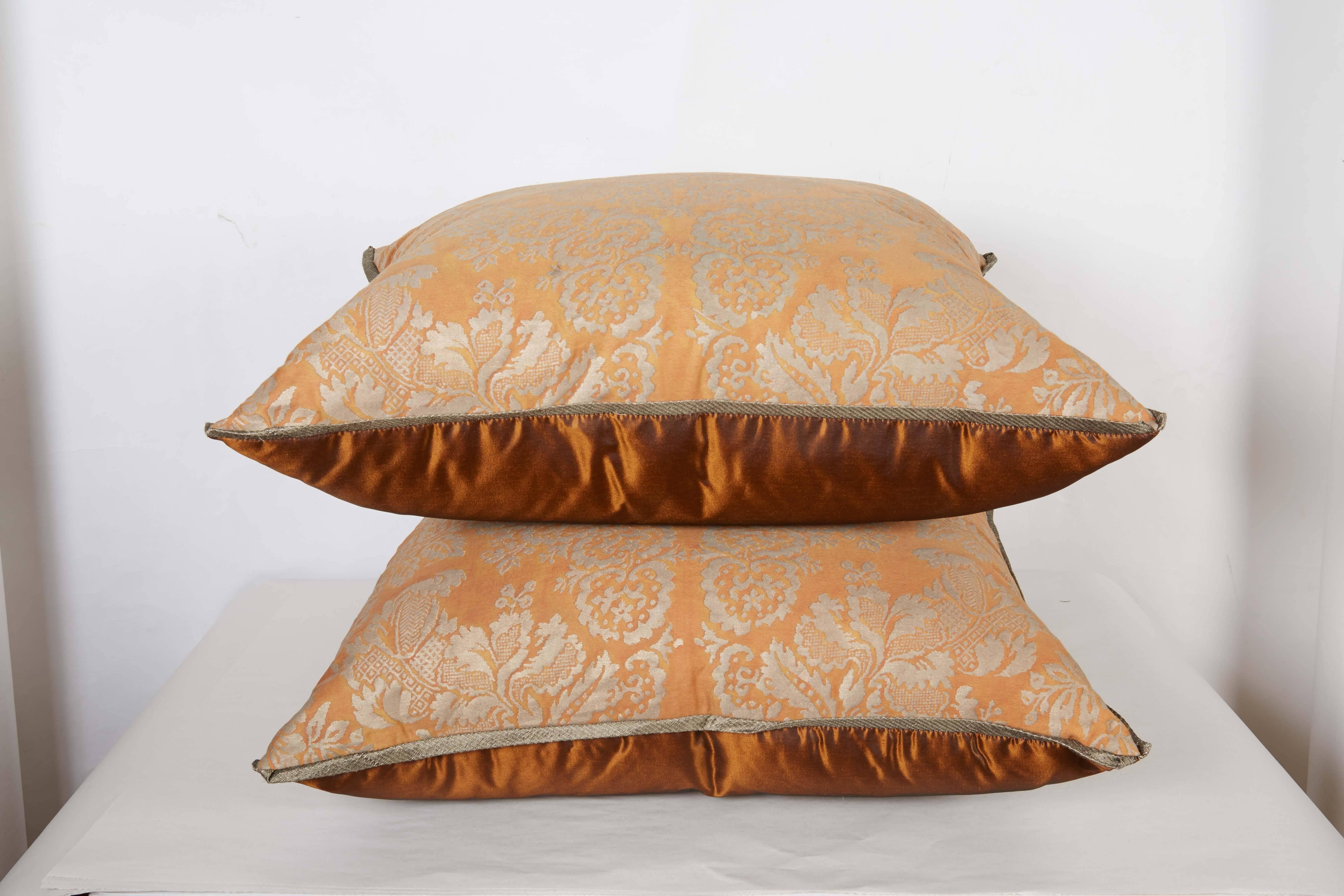 A pair of  Fortuny fabric cushions in the Solimena pattern, silvery gold and peach color way with silk bias edging and orange or rust taffeta backing, the pattern, a 17th century design named after the Neapolitan Baroque painter, Francesco Solimena
