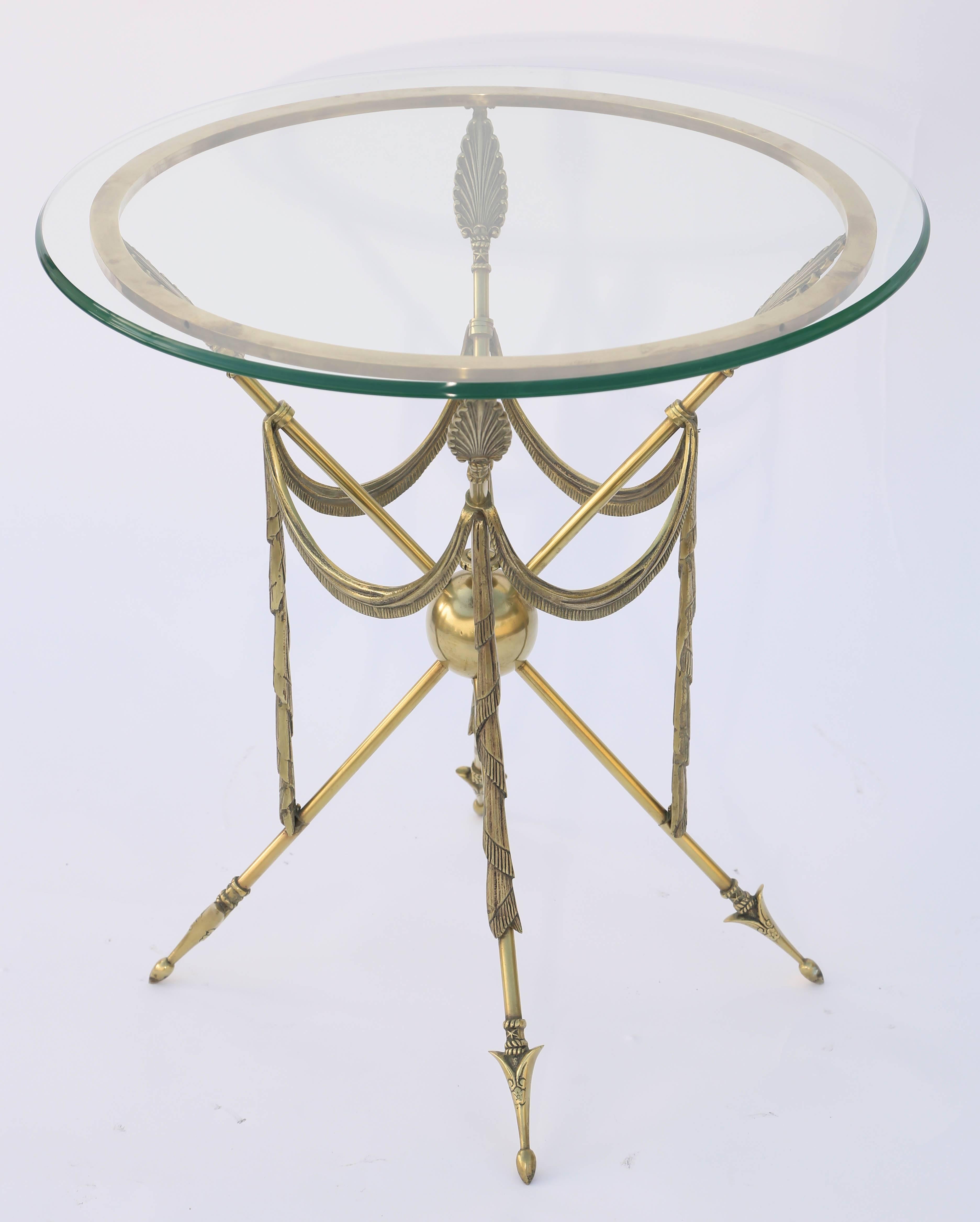 Occasional end table, of brass, having a round glass top, on conforming ring, raised on four, arrow-form, legs, draped with swagged festooning.

Stock ID: D3588