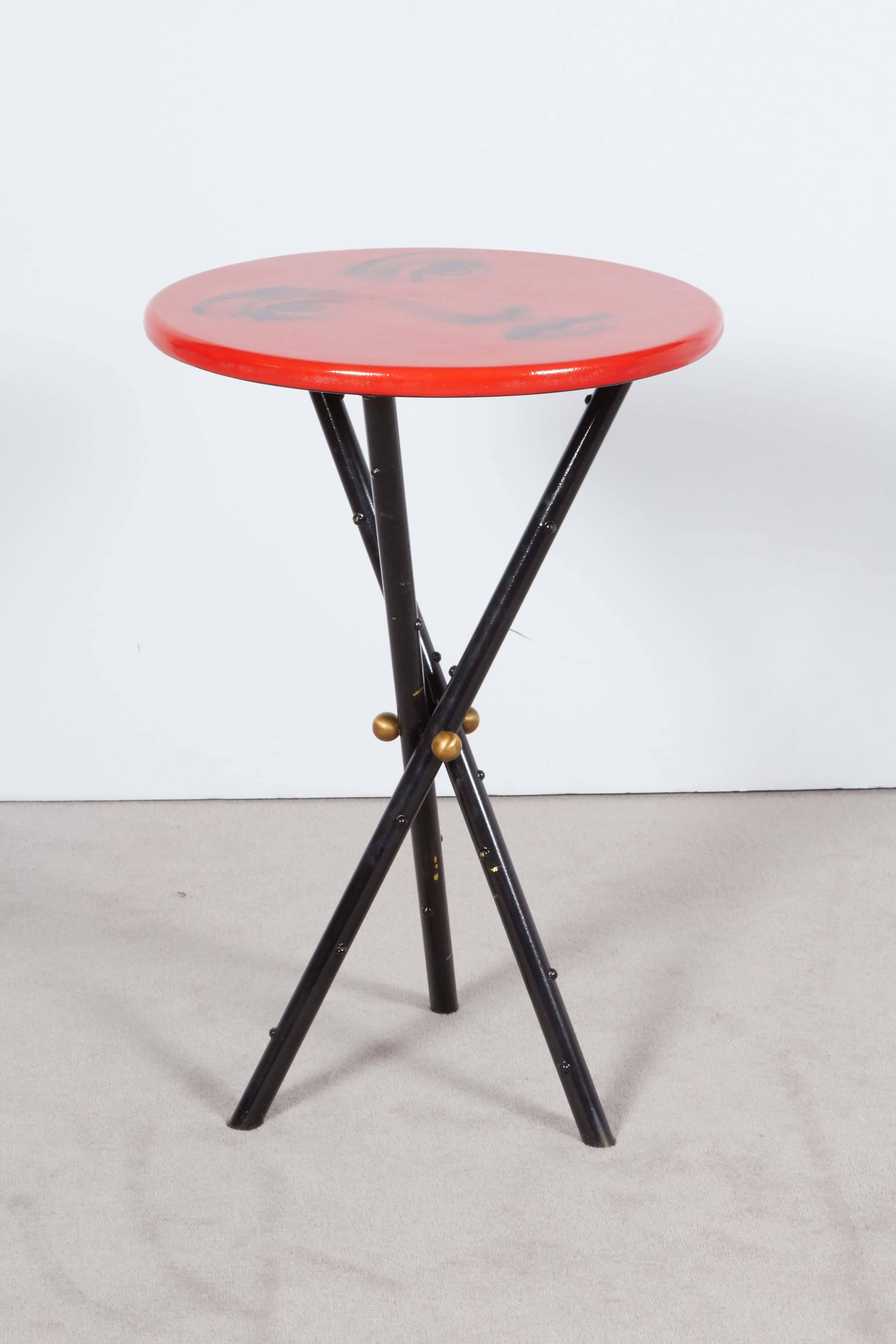 A circa 1950s side table by Italian designer Piero Fornasetti, with round red lacquered top, depicting the face of operatic soprano Lina Cavalieri, raised on crossed tripod base in black enameled brass. Markings include the original label to the