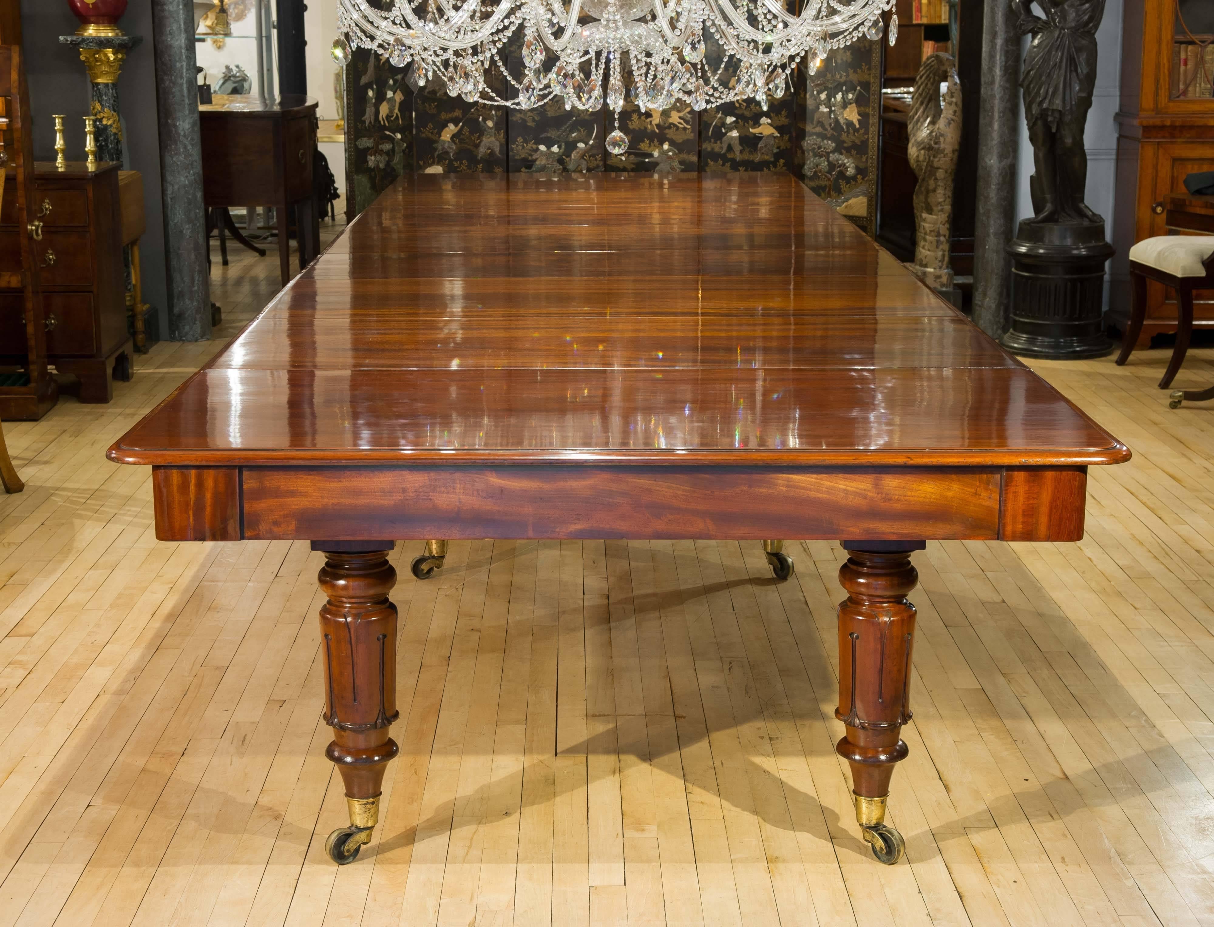 This magnificent and mammoth sized English mahogany dining table features 6 additional leaves and is supported on 8 large tulip carved legs with substantial sized brass castors. It exhibits a beautiful colouring and tone on the flame mahogany table