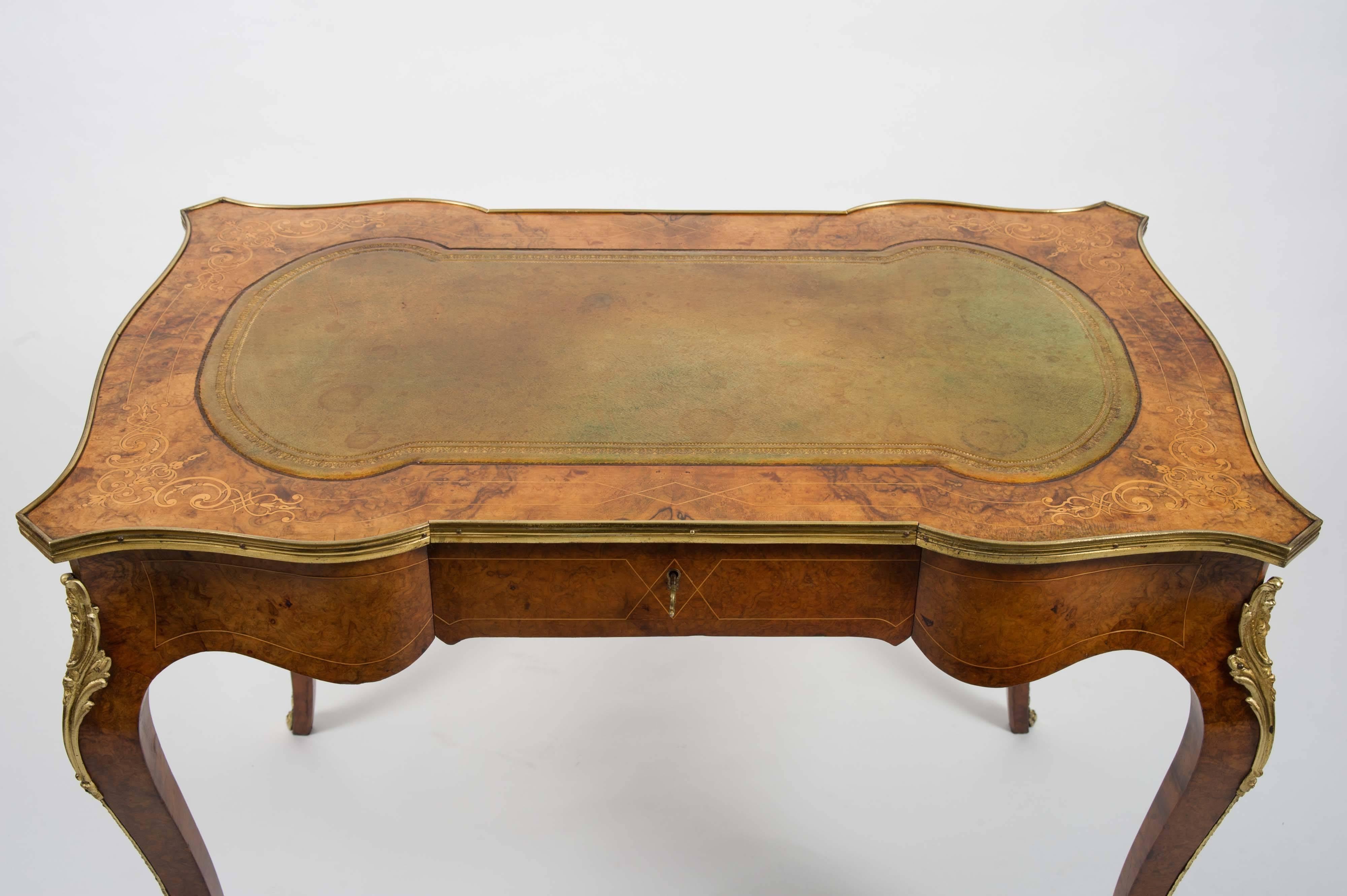 This fine quality and graceful English walnut bureau plat features a lovely inlaid design on a shaped top with ormolu edging and trim. The table has a pale green tooled leather top that follows the design shape of the table and a central locking