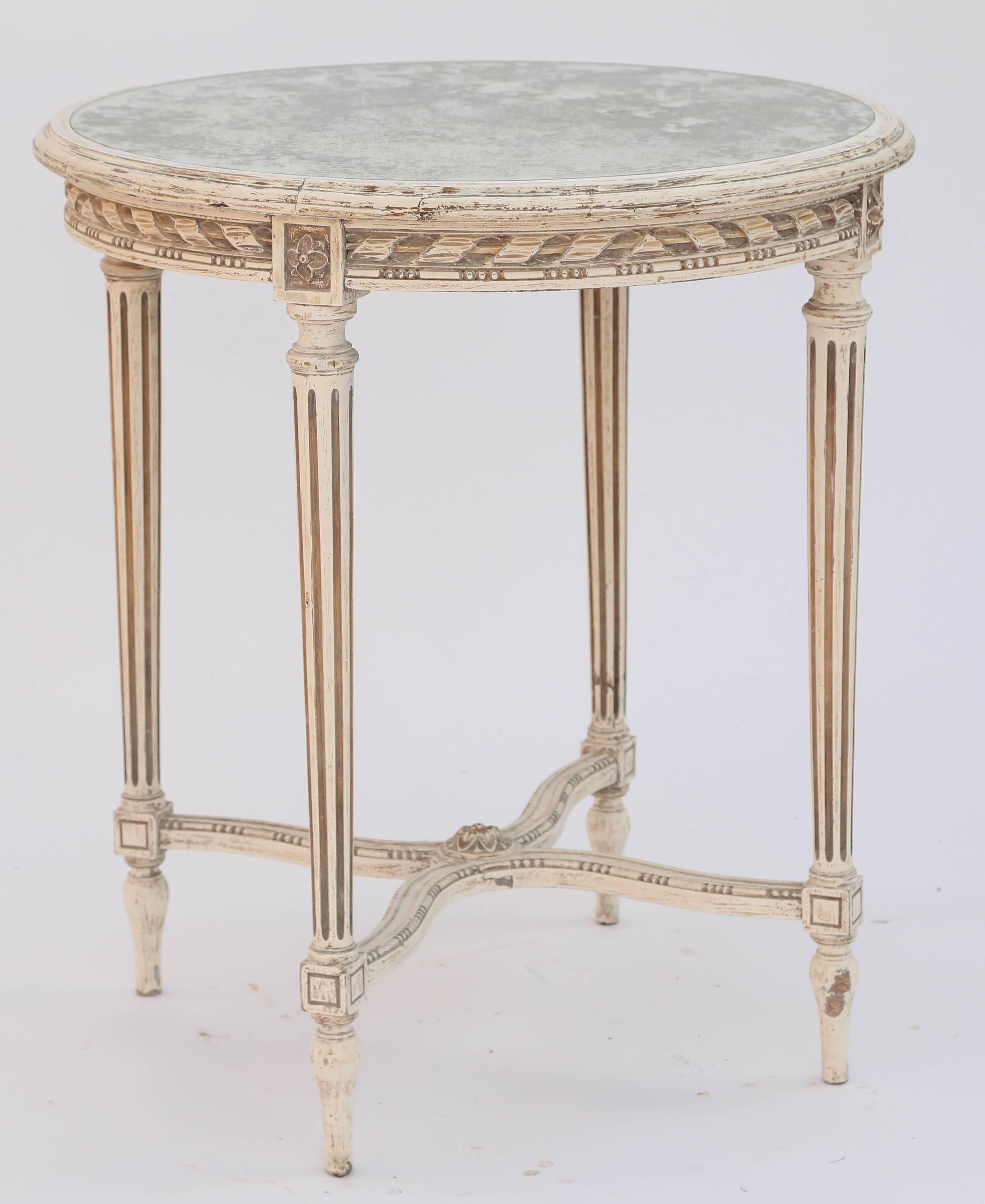 Side table, with a painted finish showing natural wear, having a round top of spotted mirror, in molded frame, its apron carved with ribbons and rods, separated by flowerhead blocks, raised on four round, tapering, fluted legs, joined by an X-form