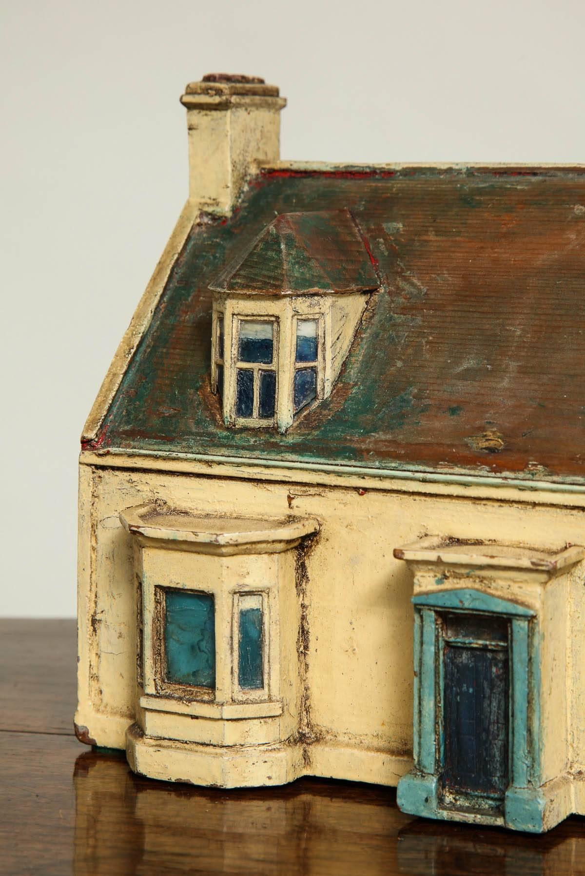 A model of a freestanding house with central hall and pedimented front door flanked by two bow fronted windows with dormer windows above and end chimney stacks. In original ochre and turquoise paint with brown painted roof and shingled dormers, with