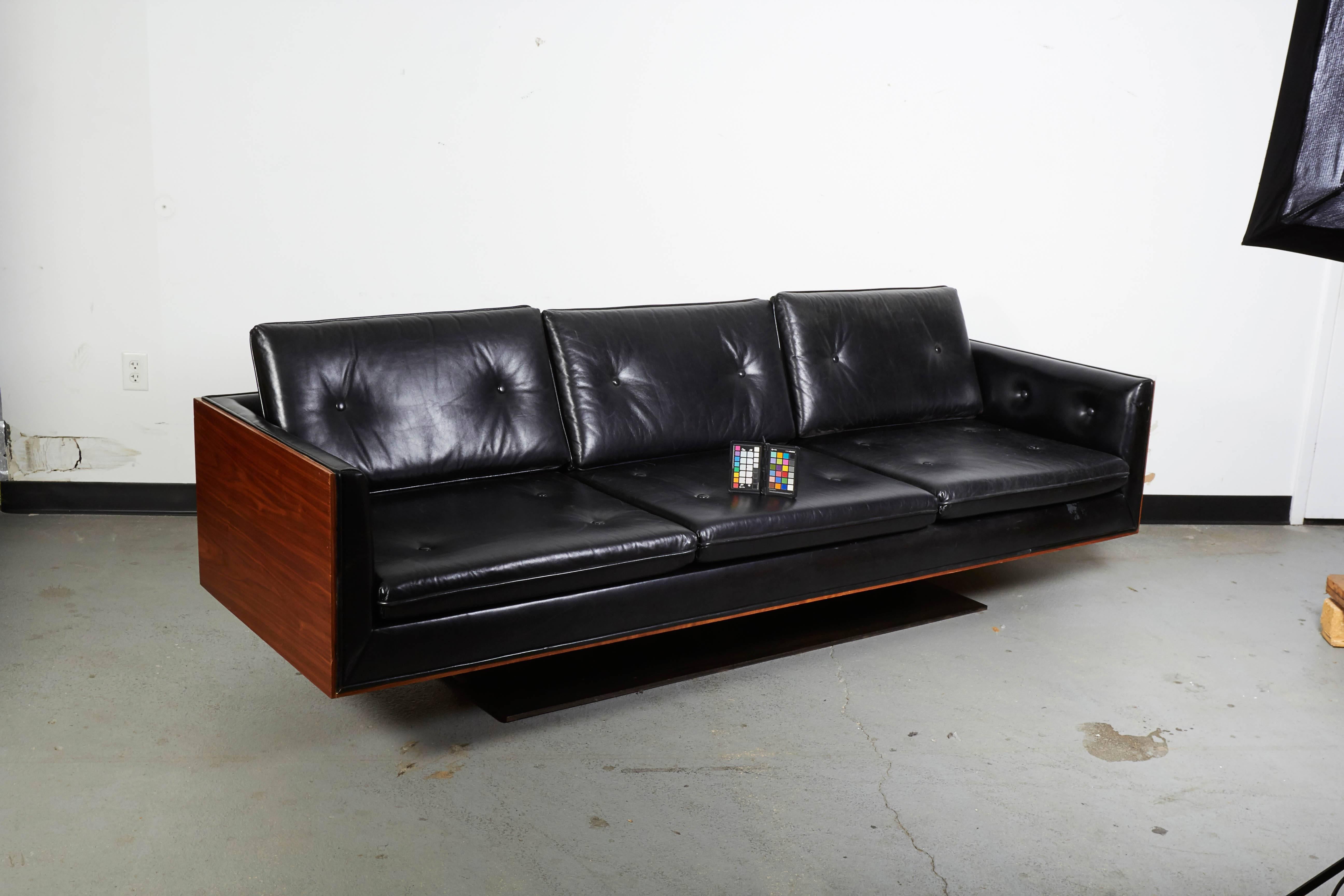 Apparently original navy blue leather upholstered sofa with tufted seat and back cushions within a bookmatched walnut veneered back and sides; raised on a steel platform to make this an early version of a floating sofa.