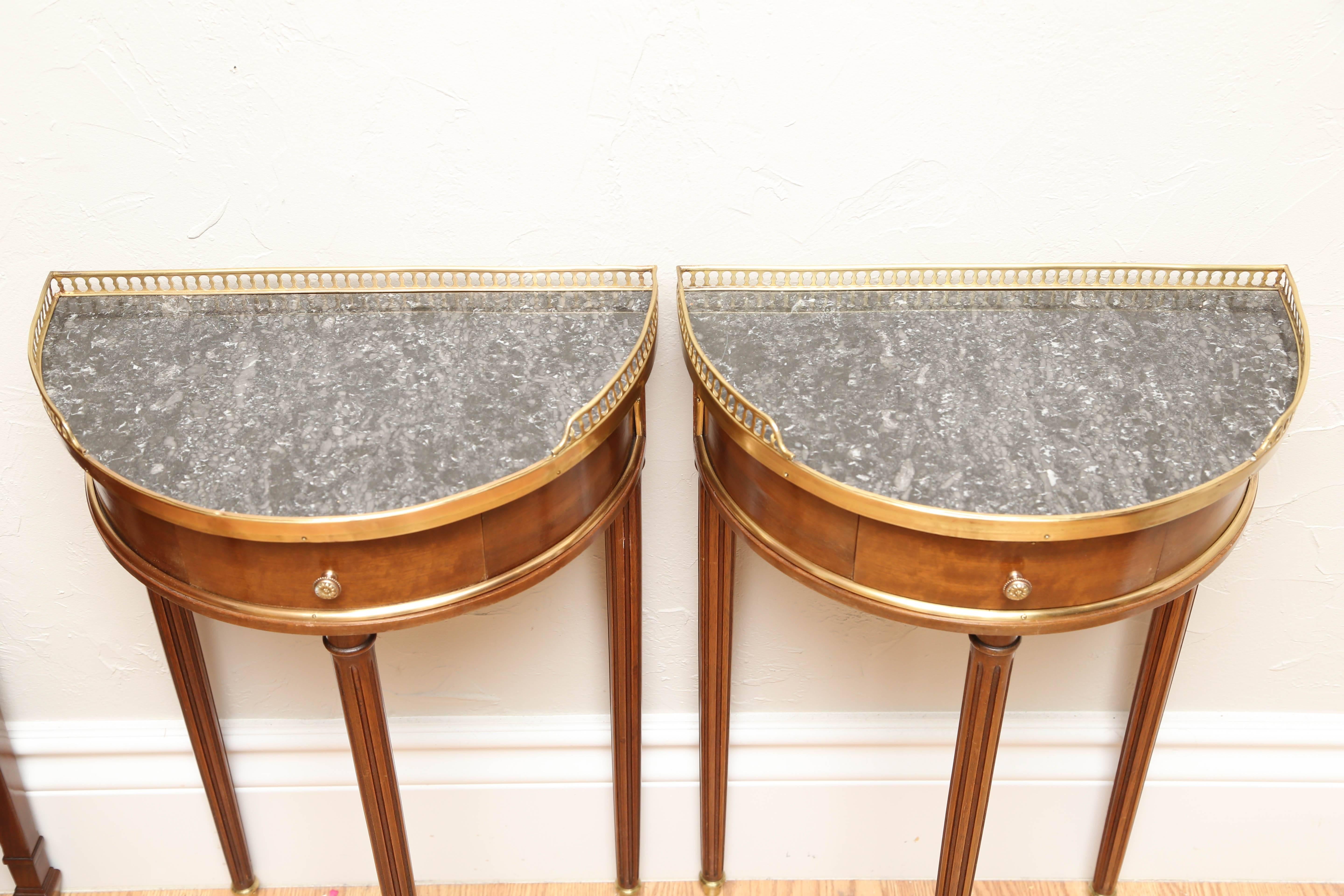 Charming pair of small French demilunes with a single drawer, gray marble-top and brass gallery.