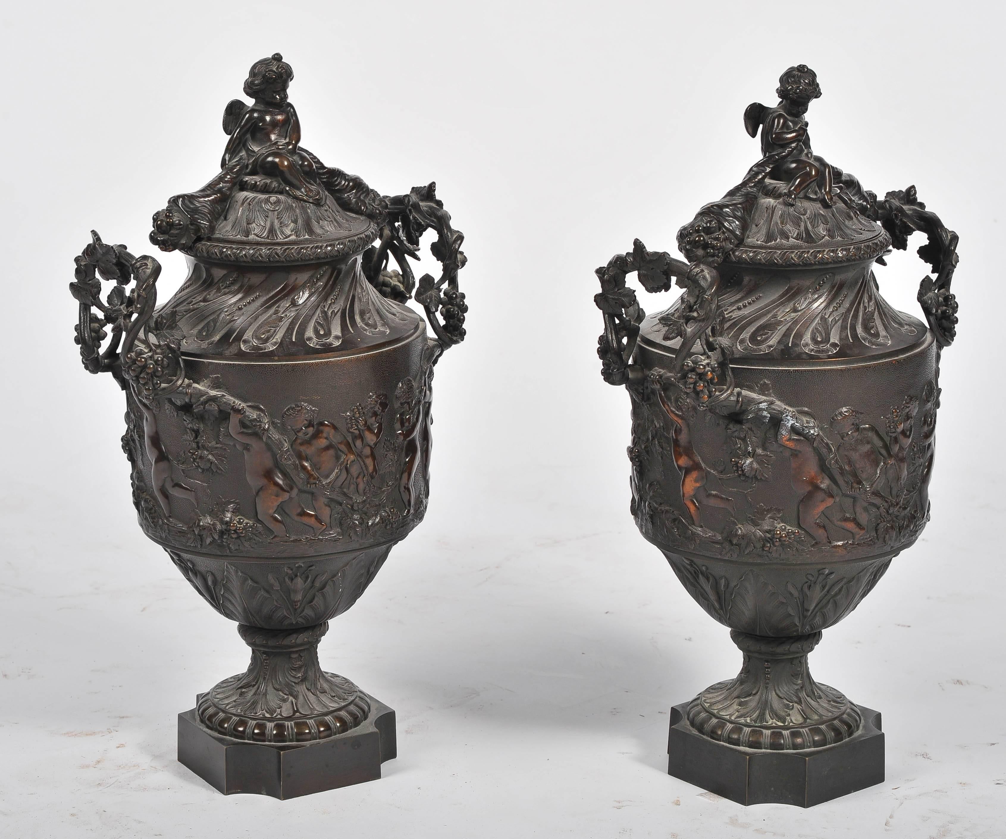 An impressive pair of 19th century Louis XVI style Bronze lidded urns, depicting Bacchanalian scenes of putti restraining a goat who is attempting to eat bunches of grapes from a vine.