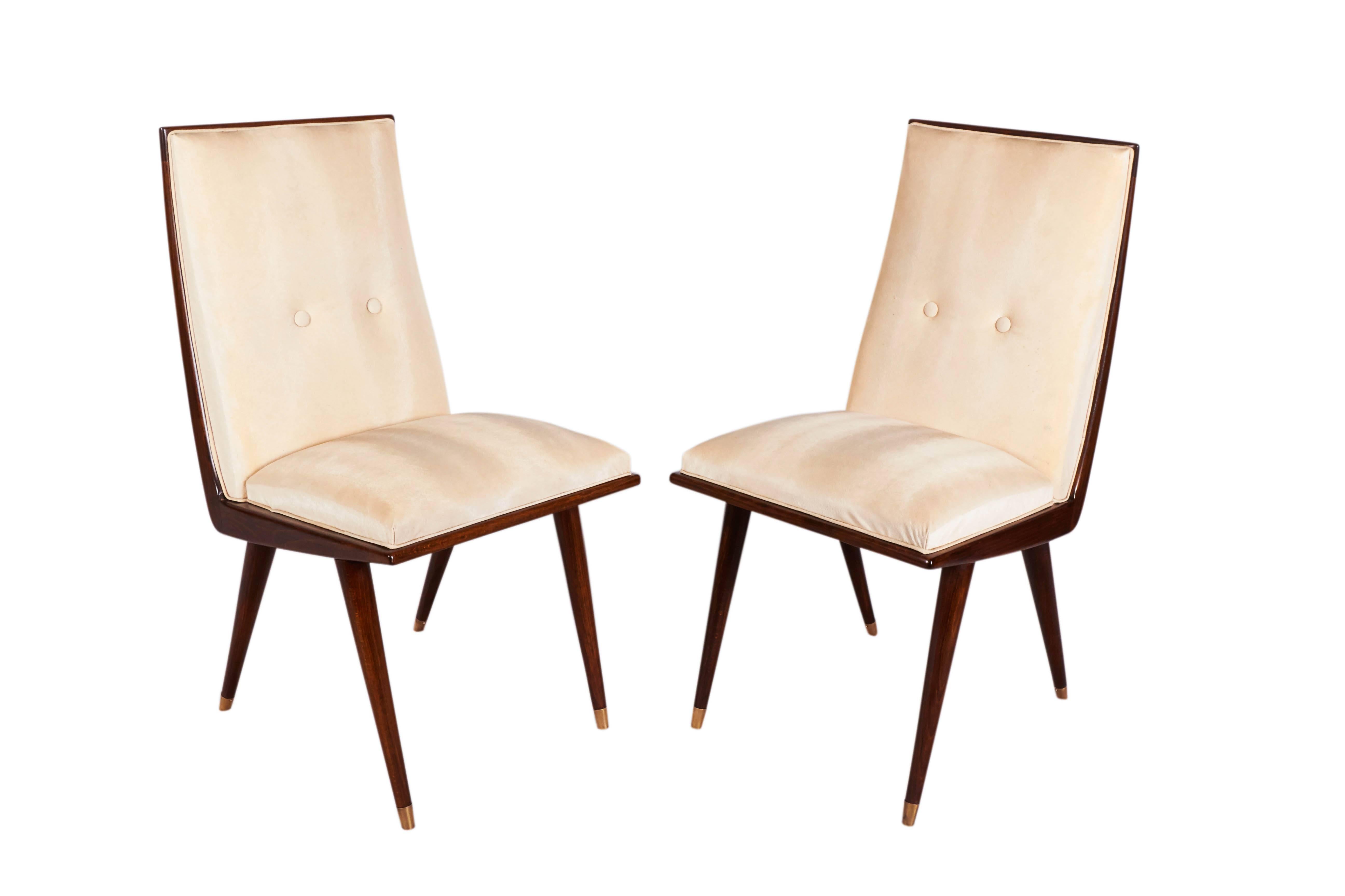A set of six French side chairs, produced in the Mid-Century Modern era circa 1950s, seats and backs upholstered in original ivory toned leatherette, against deep stained wood frames, raised on tapered legs with brass sabot feet. Very good vintage