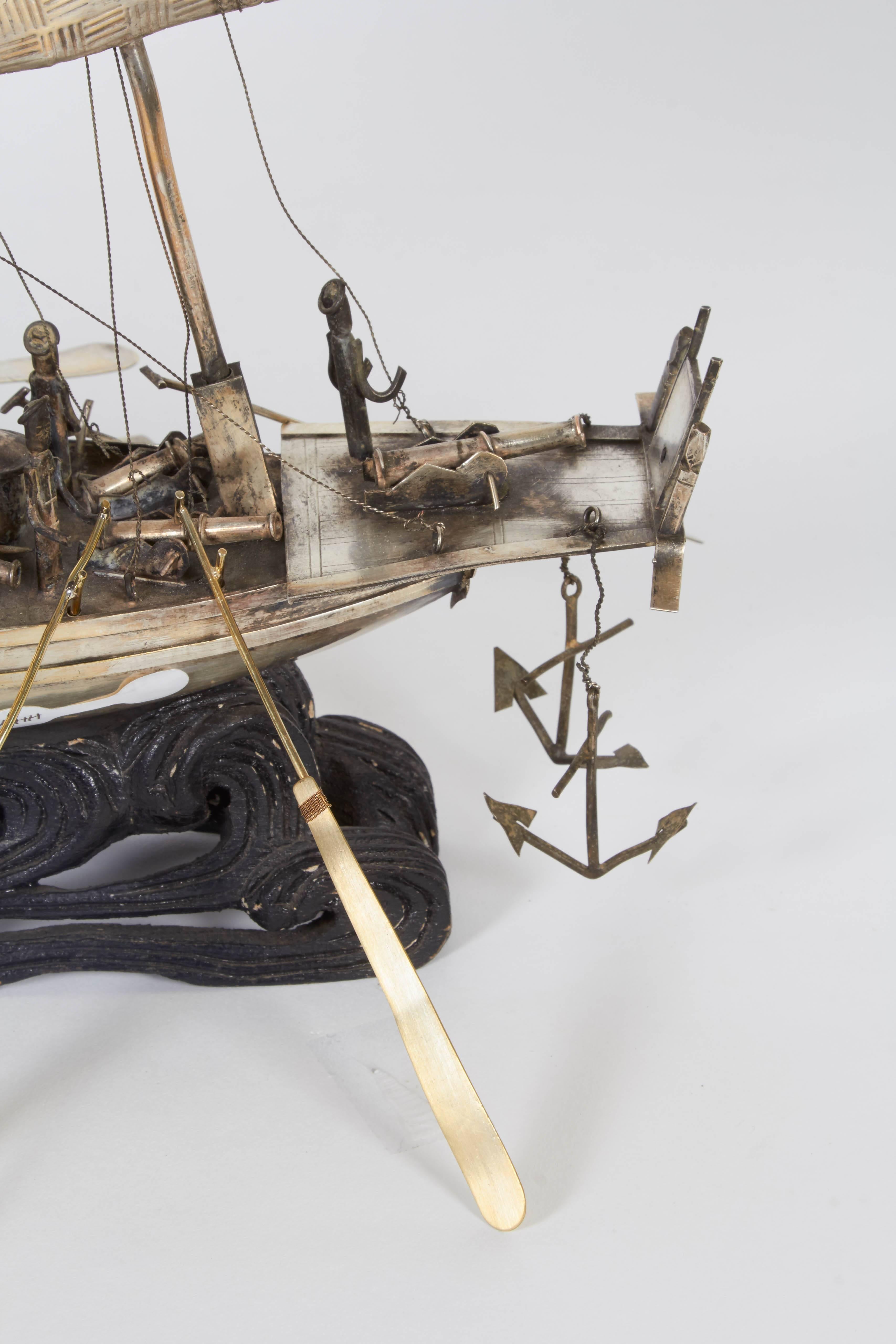 A Chinese export decorative model battleship, produced within the early 20th century period, crafted of hammered and soldered mixed metals, with seven cannons, sailors and articulating oars, mounted on a black carved wood stand, emulating crashing