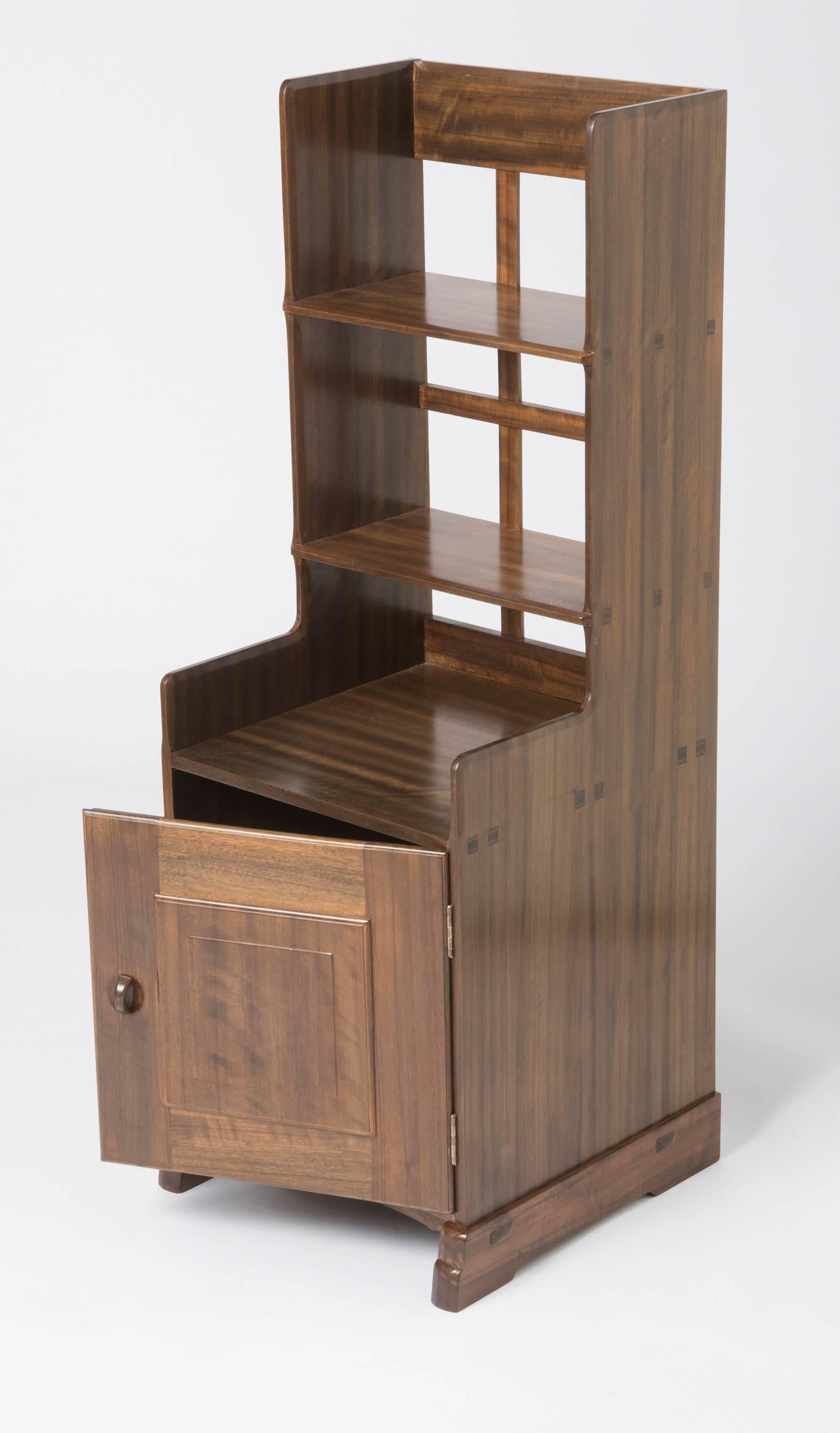 An Australian walnut bedside cabinet by Peter WaalsaAn Australian walnut bedside cabinet by Peter Waals.
The open slated back set with two shelves with chamfered edge detail.
The base with a cupboard with a stepped moulded cupboard door.
With an