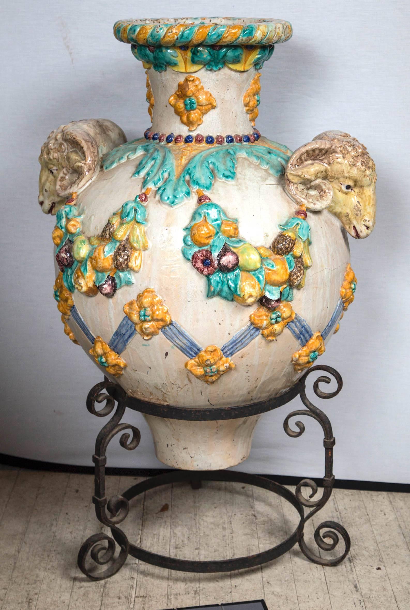 Made in Triana, Italy. This glazed urn or jar stands within a handmade iron base. Two ram's heads, garlands of fruit and stylized decoration in yellows, blues and greens, upon a cream colored body.
Underglaze blue name M. ( ?) Montalvan Triana