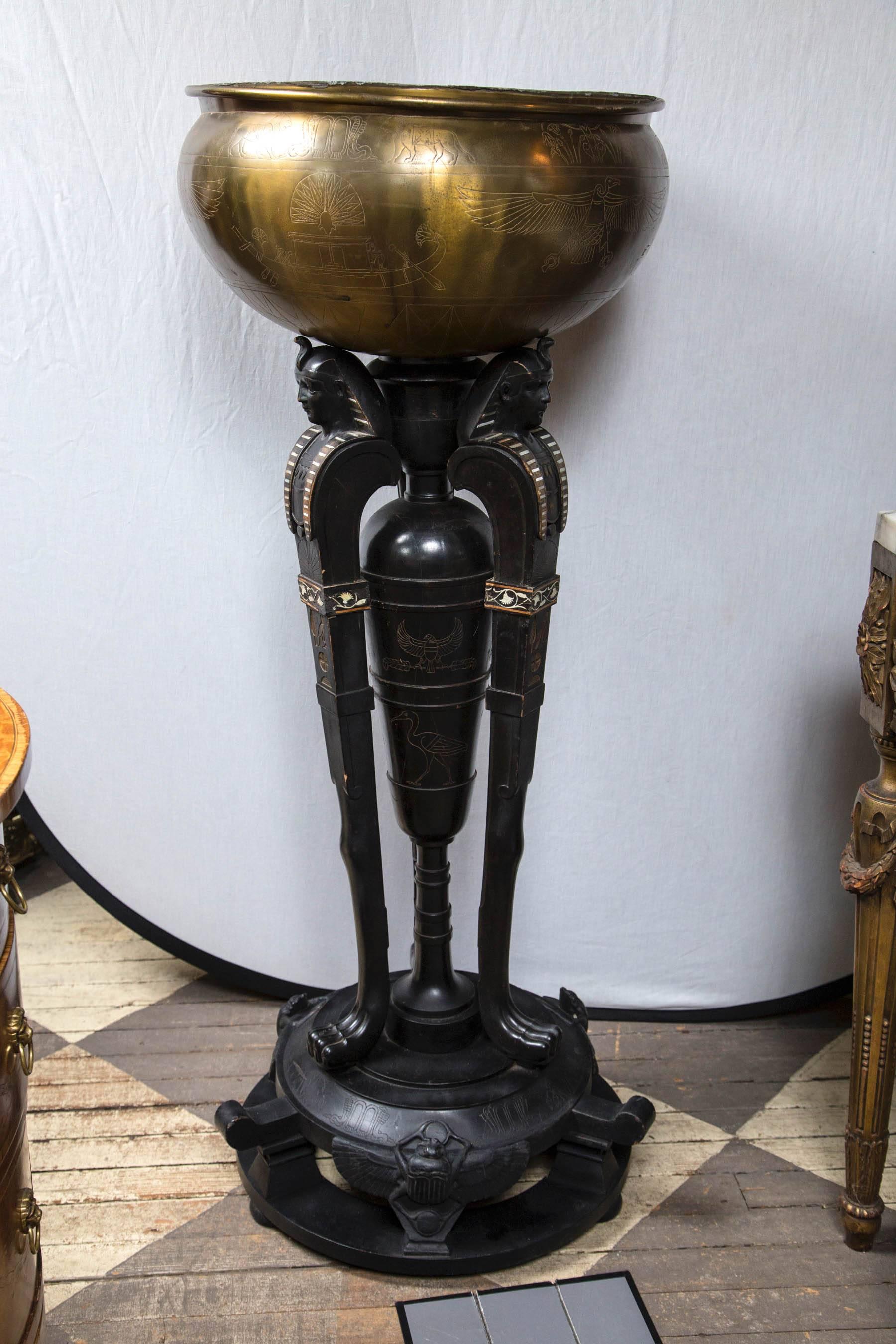 Three legs topped by Egyptian style heads and ending in four-toed animal paw feet, support a non-attached brass bowl. The heads have mother-of-pearl inlays of horizontal stripe, A few of the inlays are missing. Incised carvings highlighted in gold.