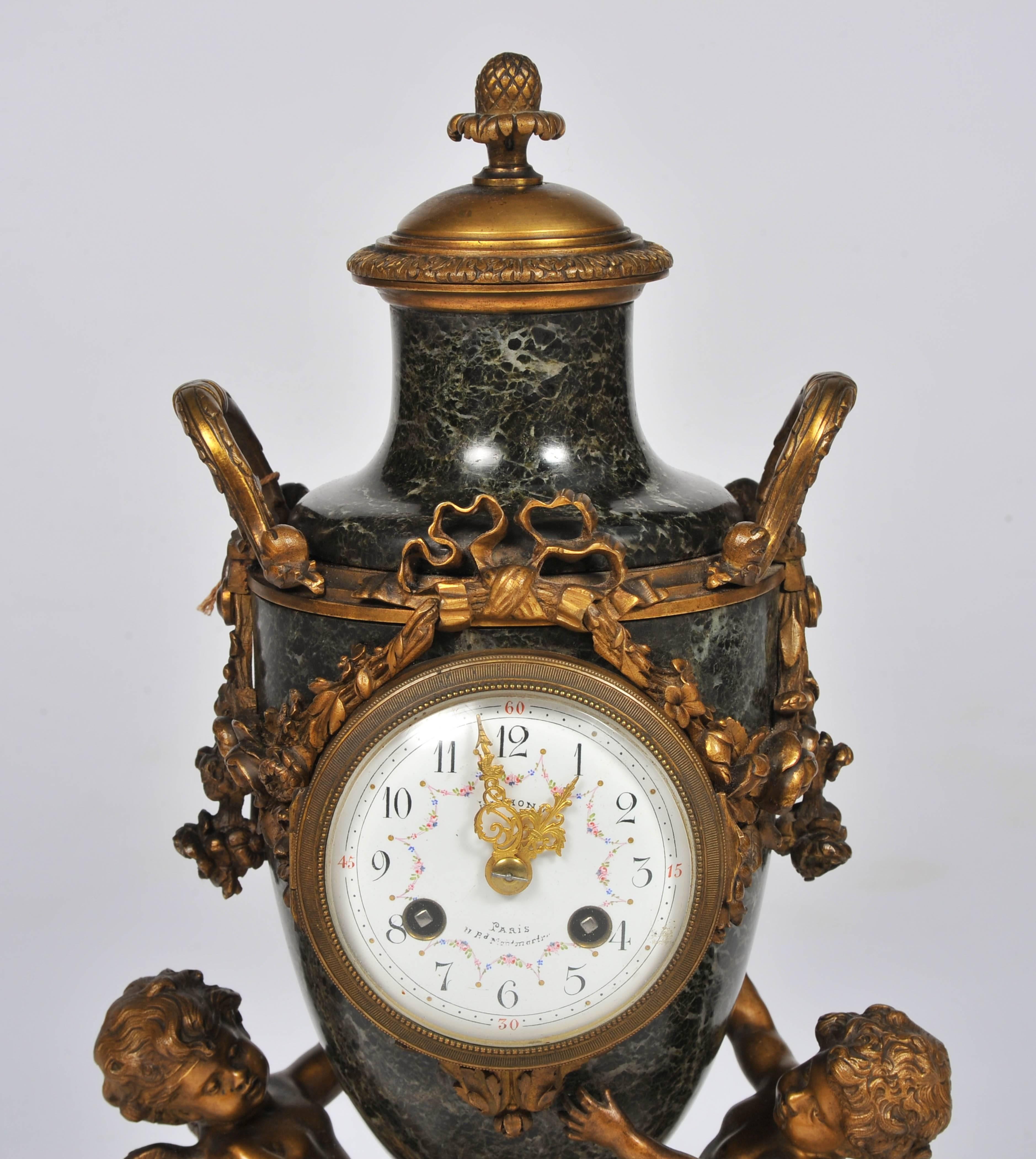 A charming French 19th century Louis XVI style gilded ormolu and green marble clock garniture, having four ormolu cherubs supporting the urn clock and two branch candelabra. The enamel dial to the clock which chimes on the hour and half hour.