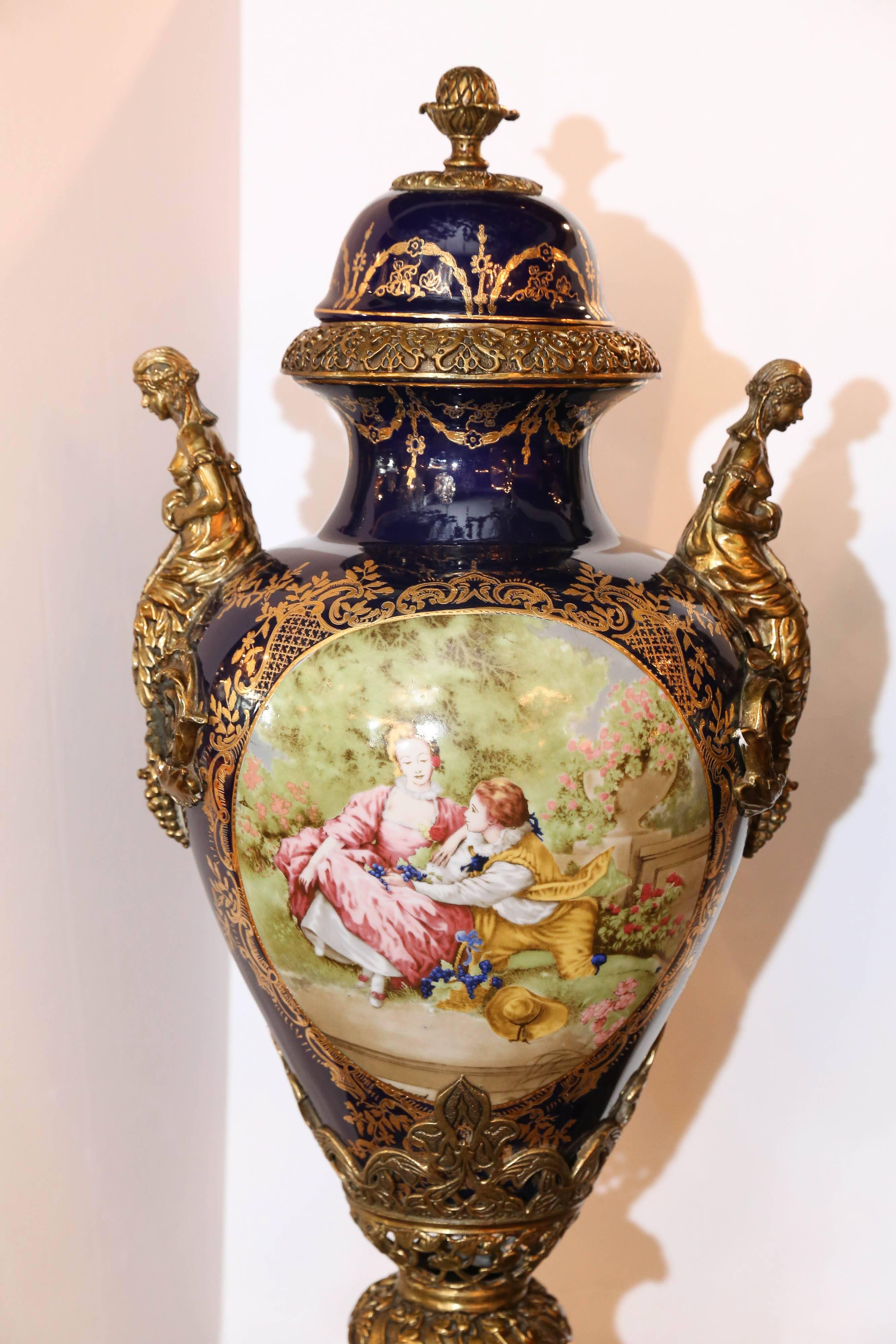 Pair of large urns decorated in cobalt blue and pale green colors
The cameo shaped front is decorated in a French garden scene
With a couple. The sides are decorated with a bronze-mounted
Woman on each side of the urn. They are mounted on a