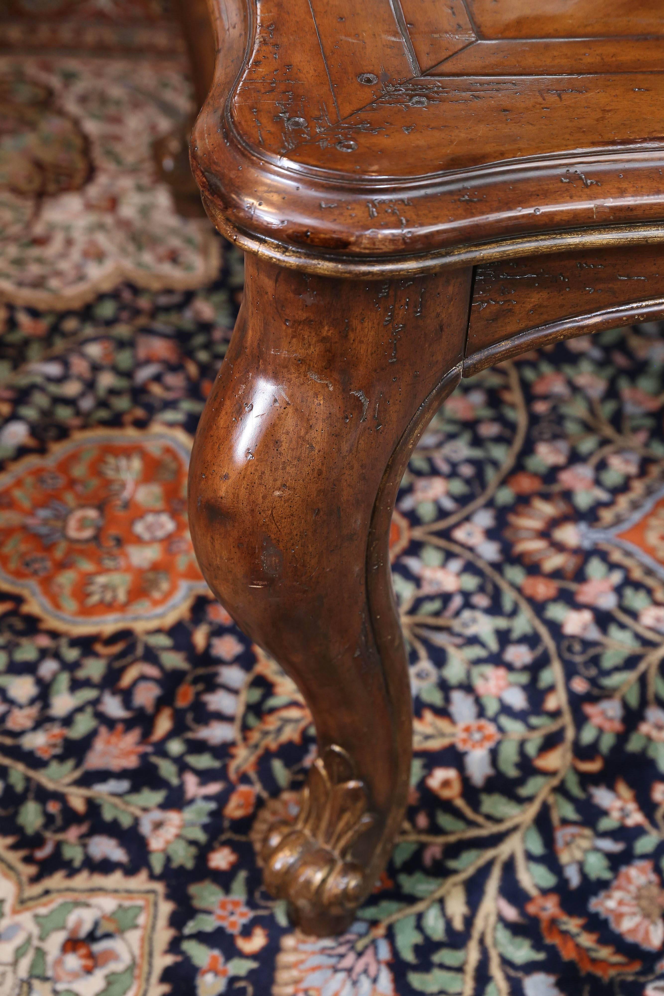 Louis XV-style table with a shaped top over an apron resting on
a large stylized curved leg; distressed walnut finish with scored
grooves. Light gilt touches decorate the table edge and the curled
foot.




 