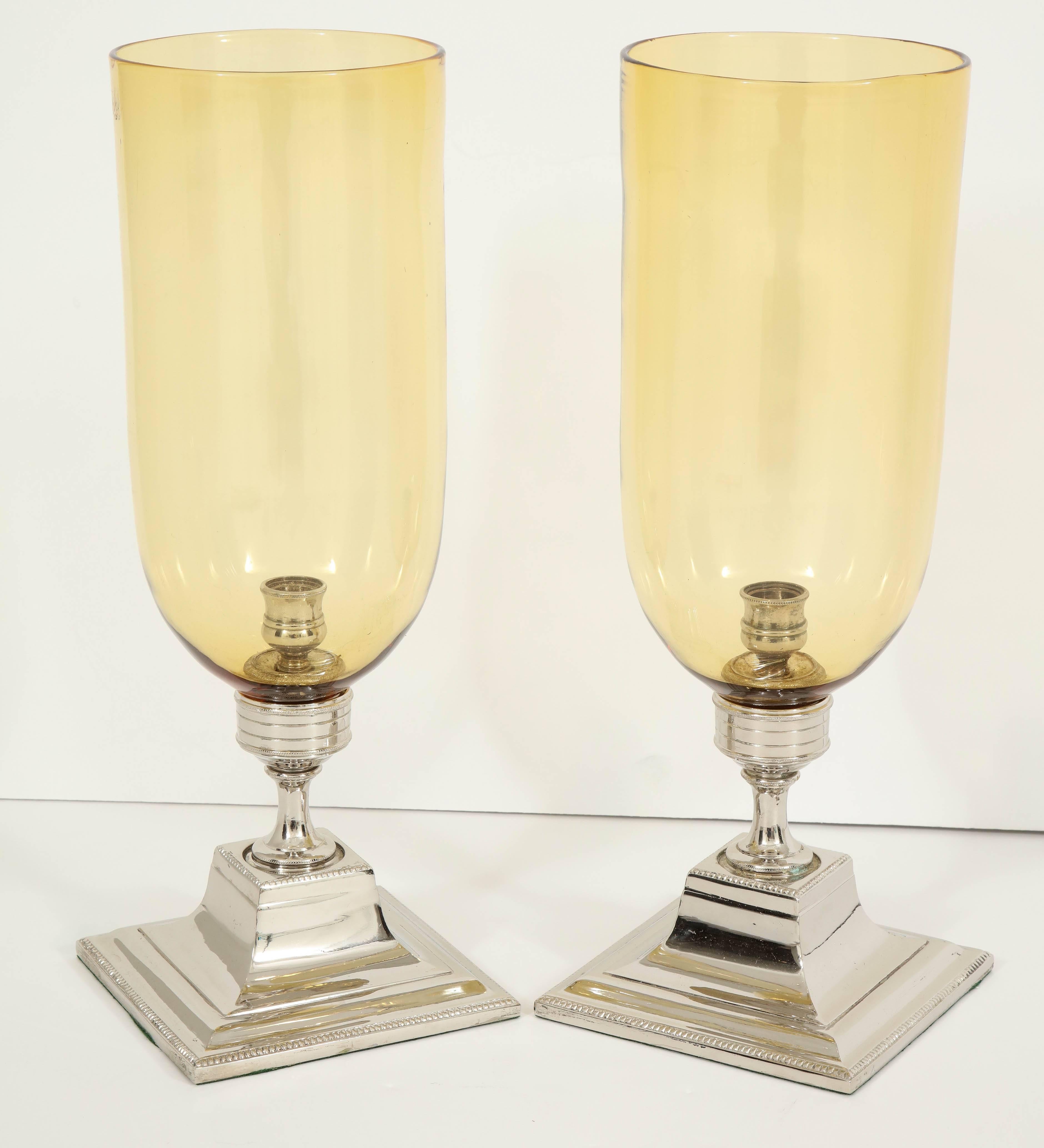 A beautiful accent for a console or dining table, this pair of amber glass hurricanes is mounted on a square, silver plate base with lovely detail.
