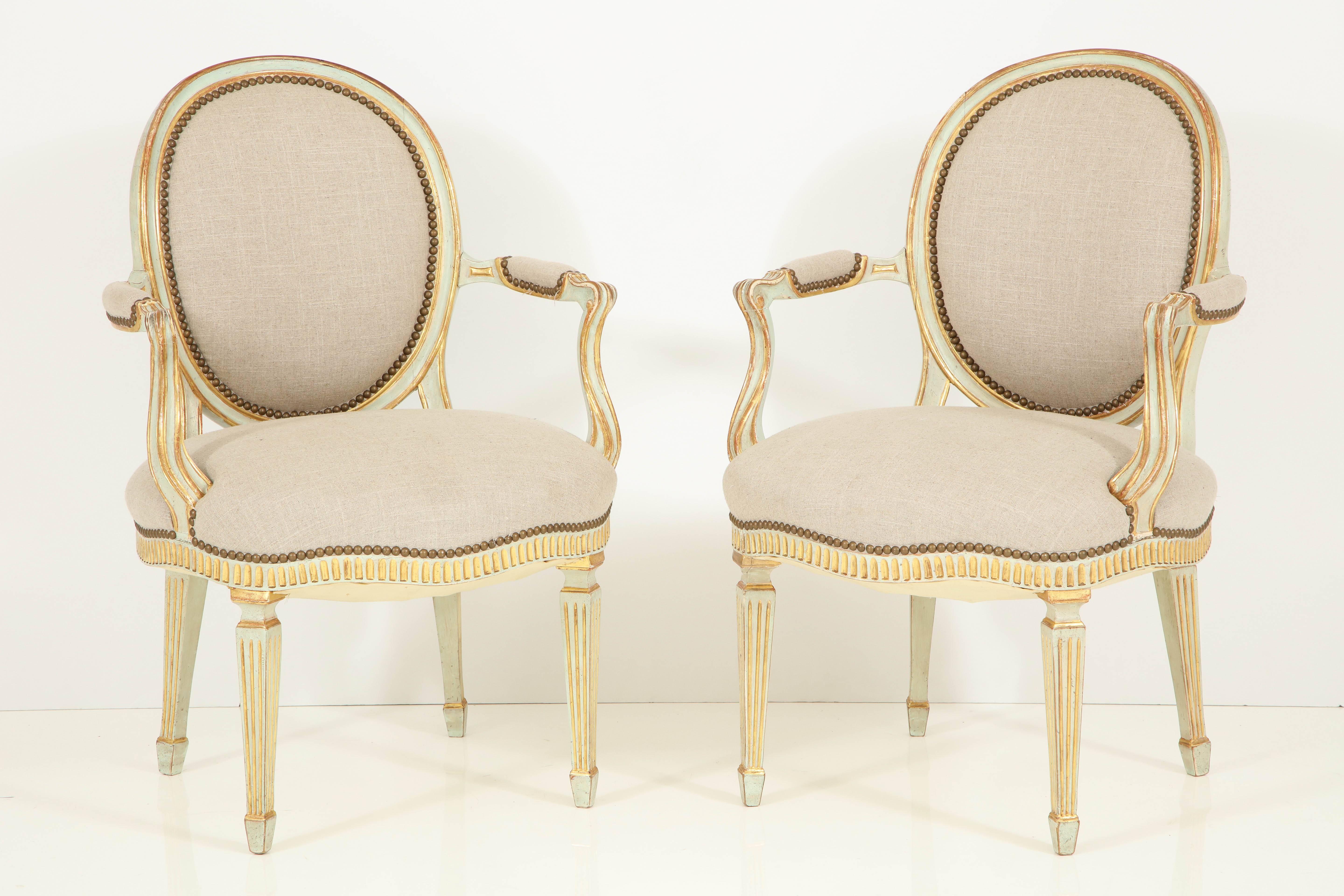 A beautifully crafted pair of Louis XVI Style fauteuils with oval backs, a serpentine front, a fluted apron and fluted, slightly tapered legs. The painted finish is a creamy green with gold leaf accents. The chairs are newly re-upholstered in a