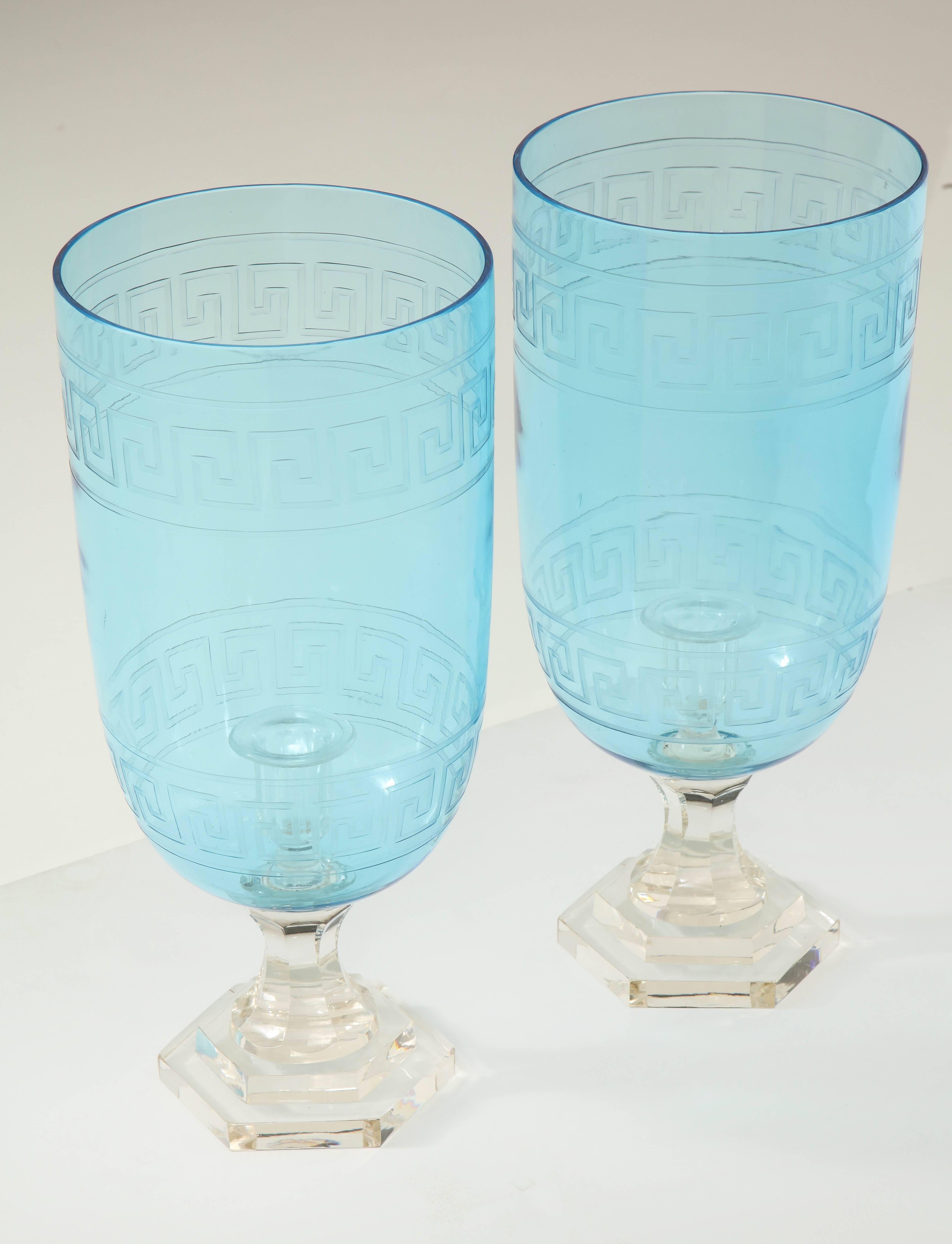 A pair of glass hurricane candleholders in a stunning blue with an etched Greek key design on a clear glass pedestal. Perfect on a console, mantel or centrepiece.