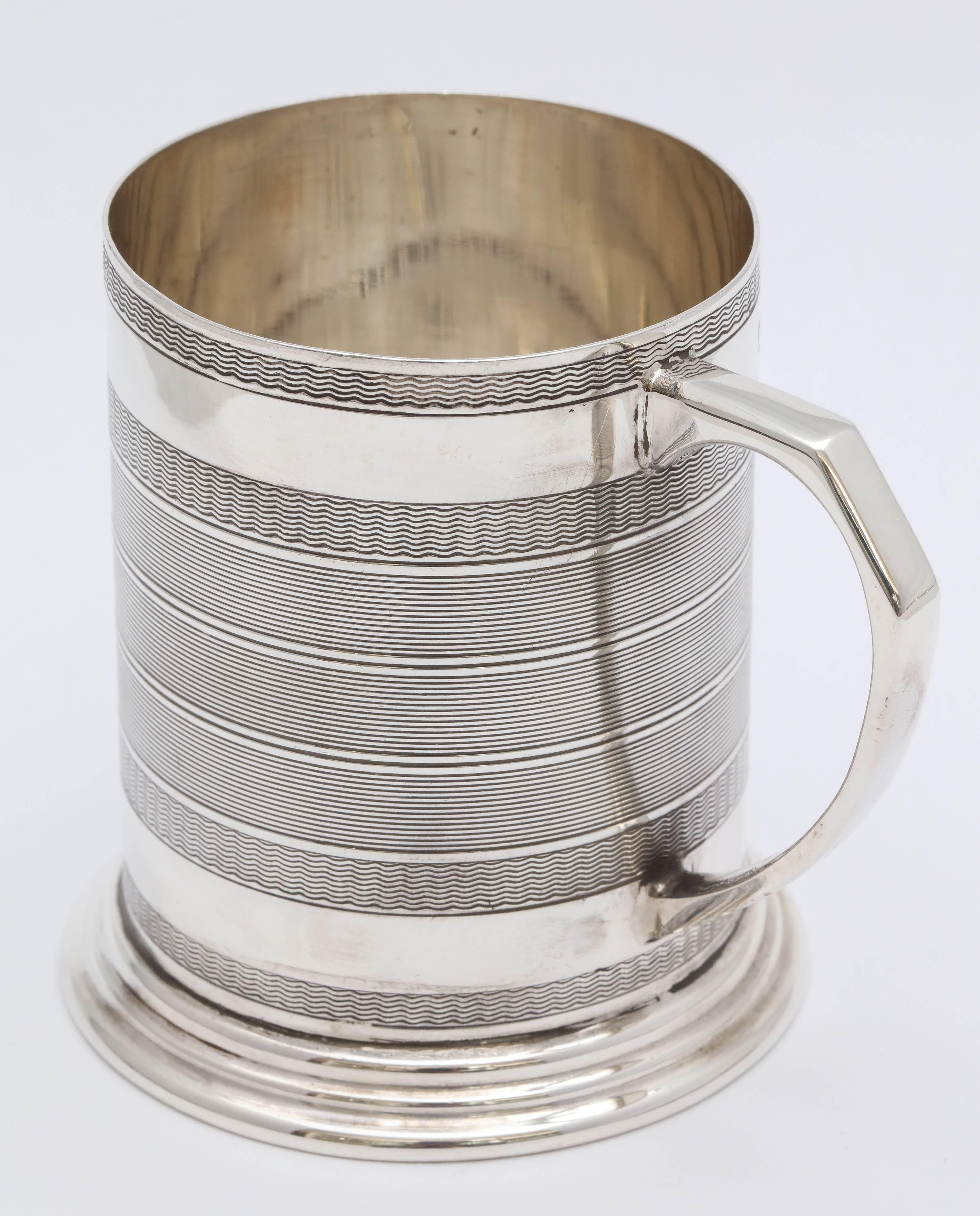 Sterling silver baby mug in the Edwardian style, Birmingham, England, 1944, Hukin & Heath, makers. Engine, turned design; stepped up base. Measures: 2 3/4 inches high x 2 1/2 inches diameter x 3 1/4 inches across, rim to handle. Weighs 2.875 troy