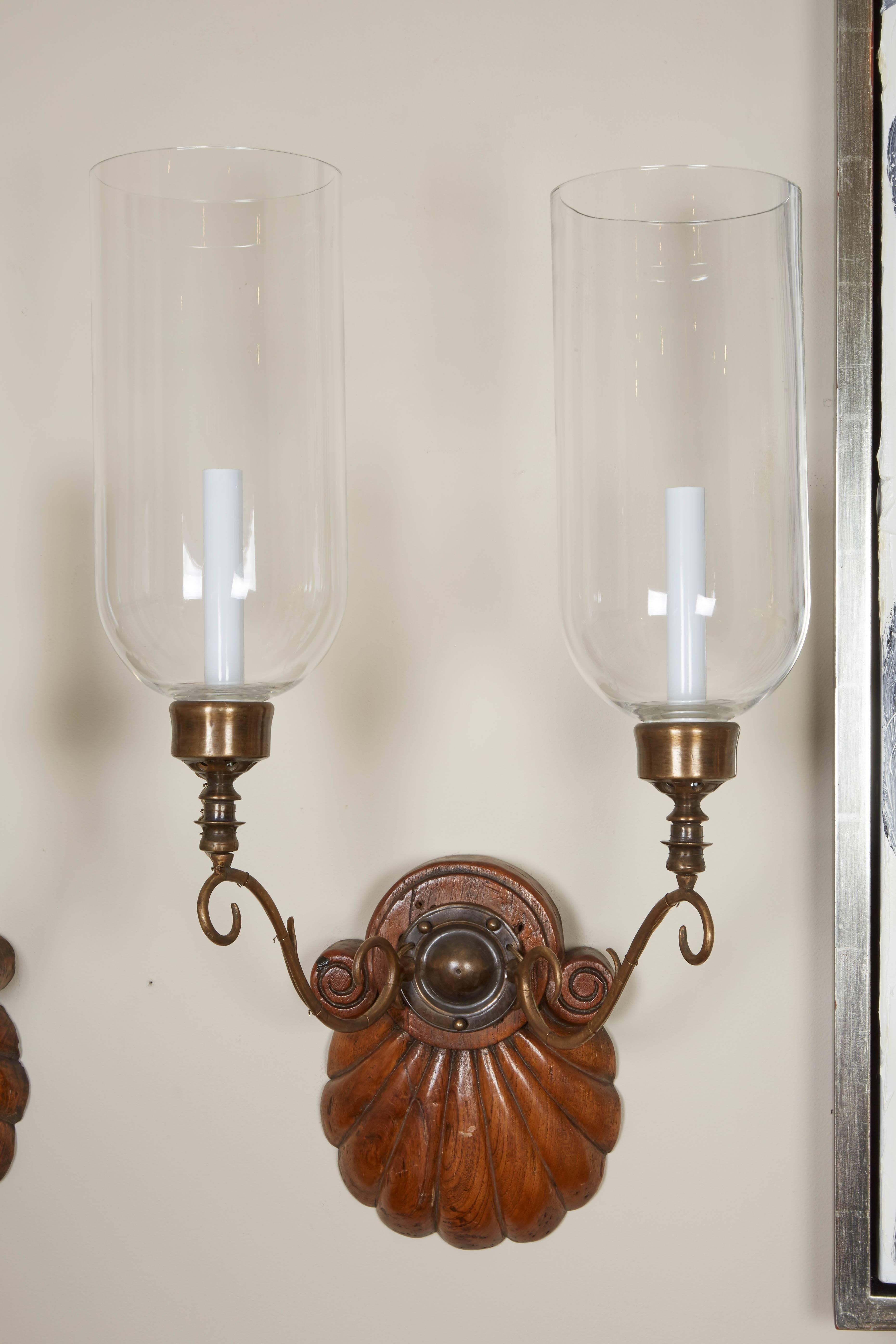 A pair of Anglo-Indian two-light sconces, hardwood shell form backplate issuing bronze out-scrolled candle-arms supporting a single Edison socket with hurricane shade, circa 1890.
Provenance: These sconces were purchased by Albert Nestle in India,