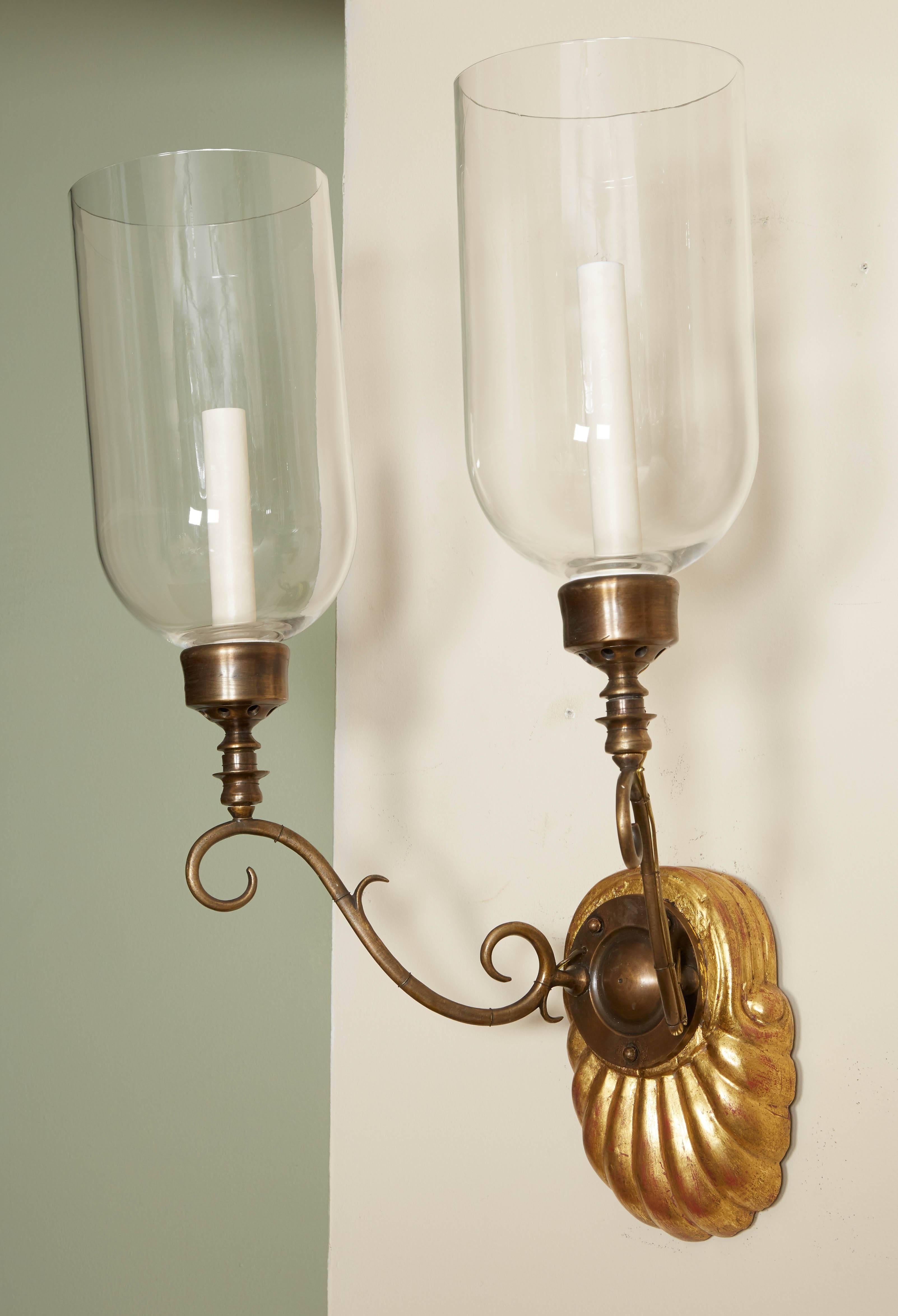 A pair of Anglo-Indian two-light sconces, gilded hardwood shell form backplate issuing bronze out-scrolled candle-arms supporting a single Edison socket with hurricane shade, circa 1890.

Dimensions; 
24
