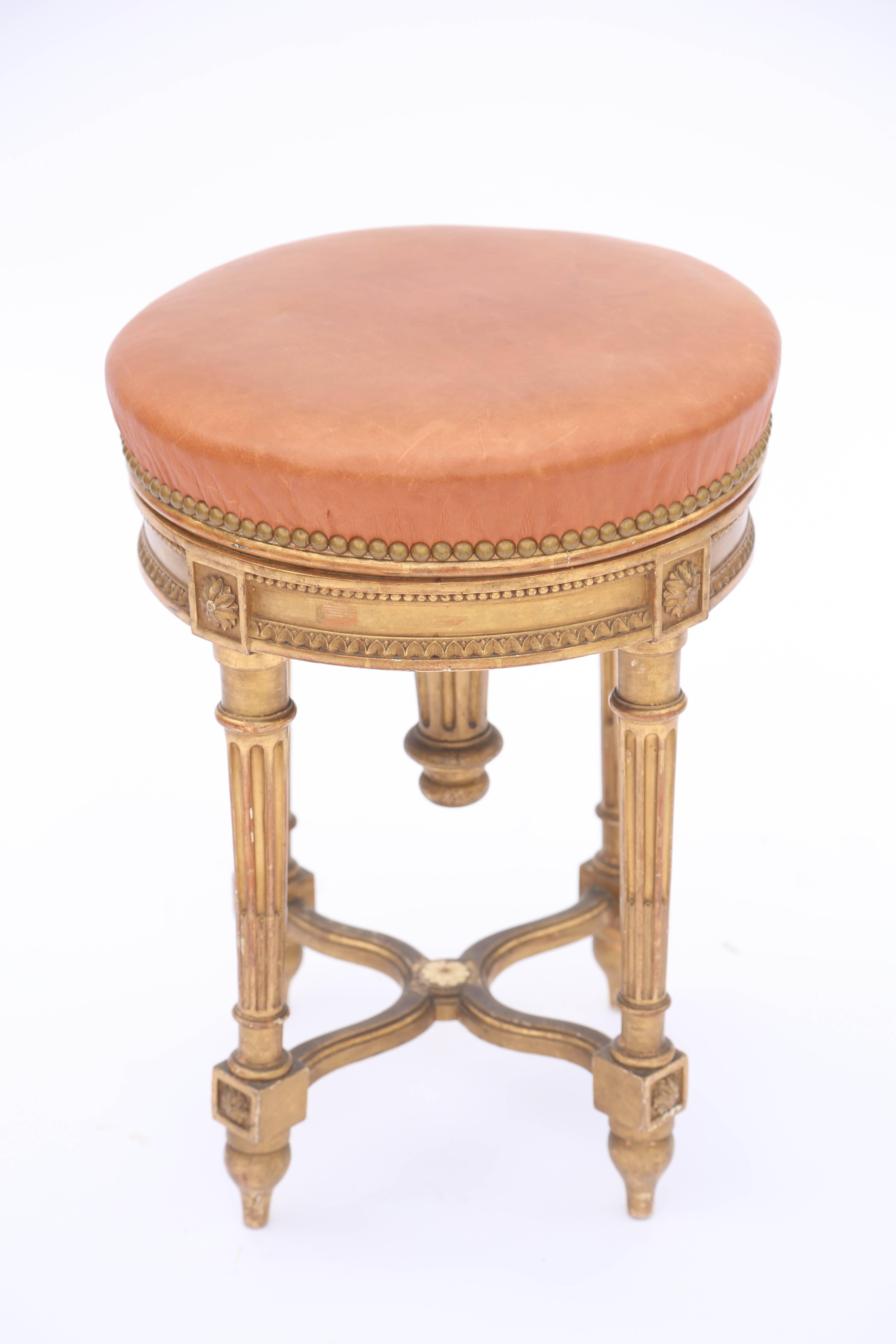 Adjustable height piano stool, in the Louis XVI style, of carved giltwood, its rotating round seat upholstered in leather with nailheads, on fielded apron carved with cockbeading and gadrooning, raised on four round, tapering, fluted legs, joined by