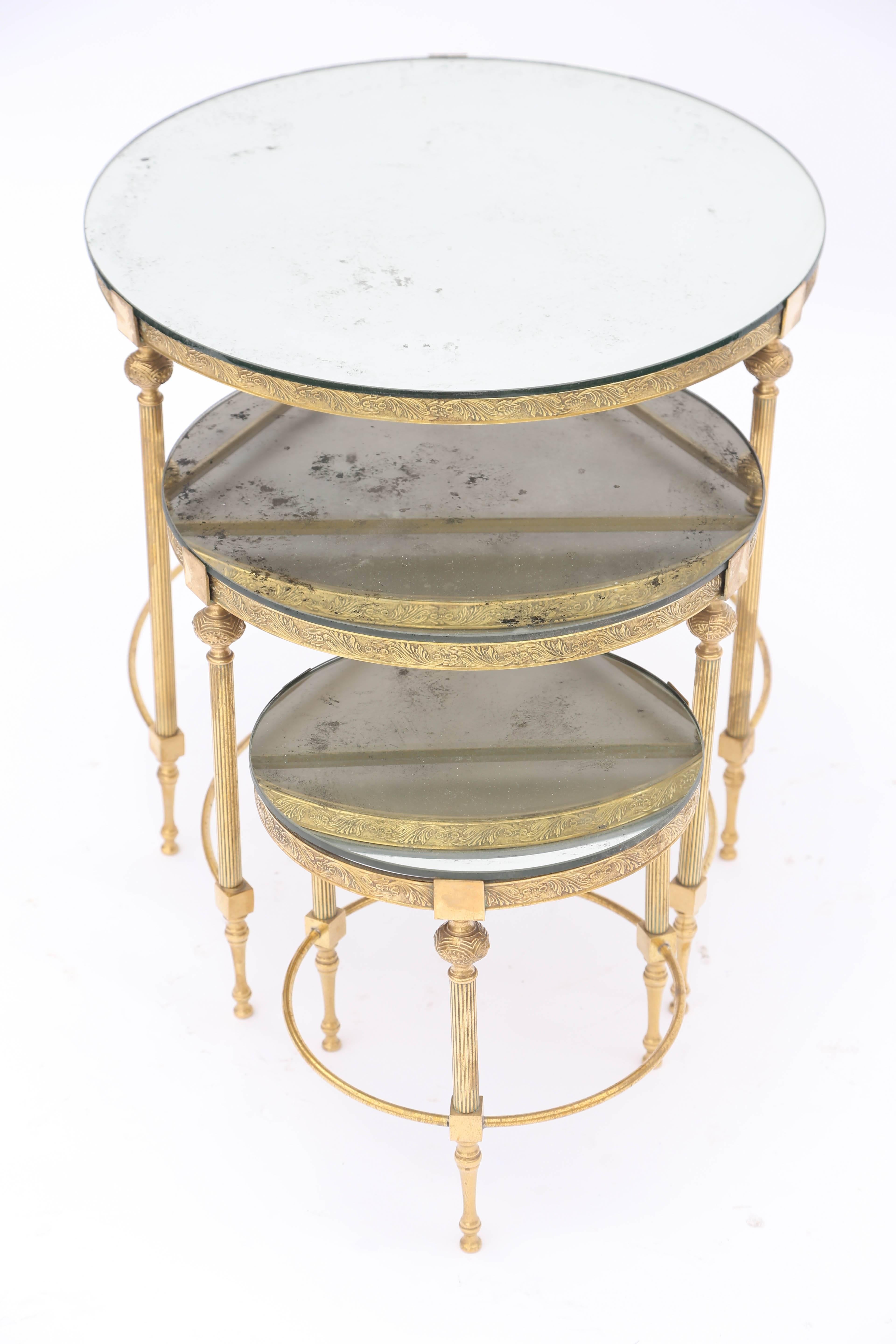 Unusual set of three stackable accent tables, in the manner of Bagues, in graduating size, each having a round top of aged mirror, on gilt metal base, with apron decorated by foliate scrollwork, raised on round, fluted legs, each with a knopped